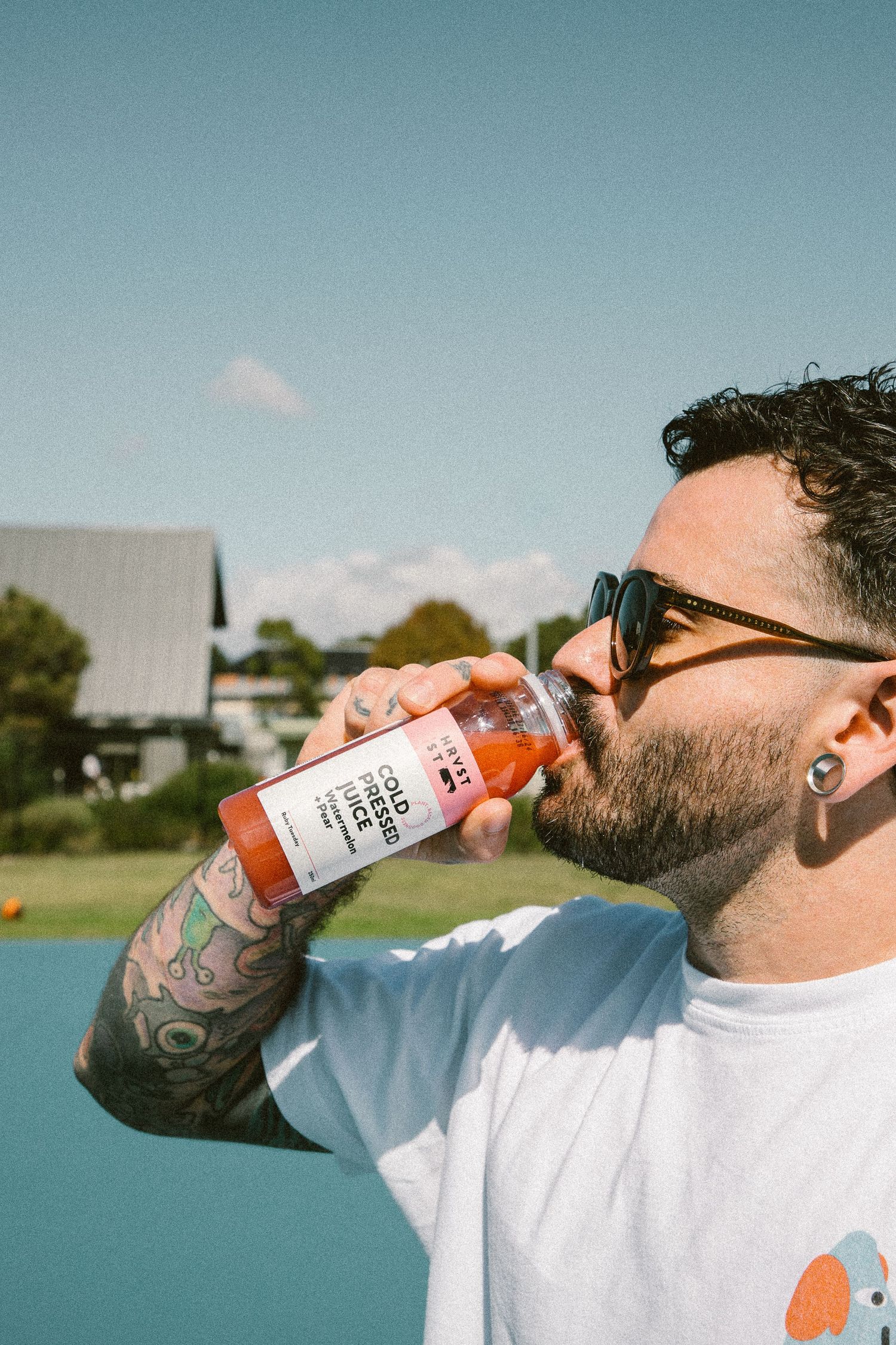 Tattooed man with sunglasses drinking a cold pressed juice bottle outdoors on sunny day.