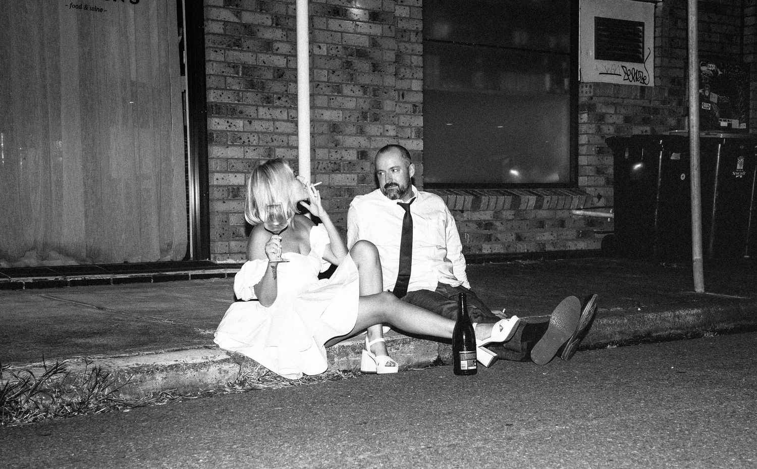 Black and white photo of bride and groom sitting on curb outside brick building after wedding reception.