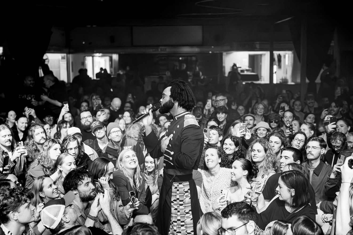 Black and white photo of a guitarist performing on stage surrounded by an enthusiastic crowd at a live music venue.