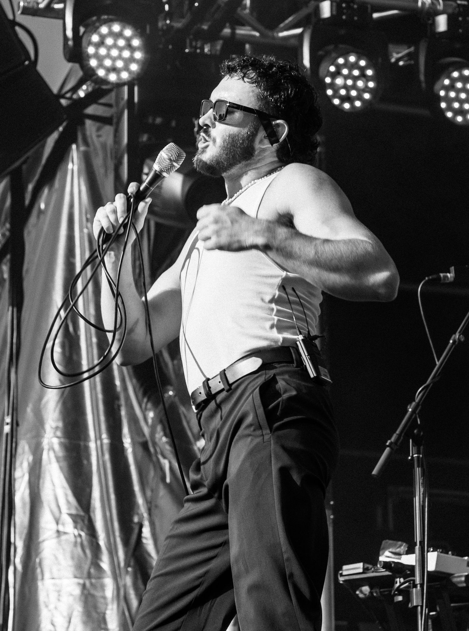 Black and white dramatic shot of male performer of San Cisco in tank top and sunglasses singing on stage with microphone.