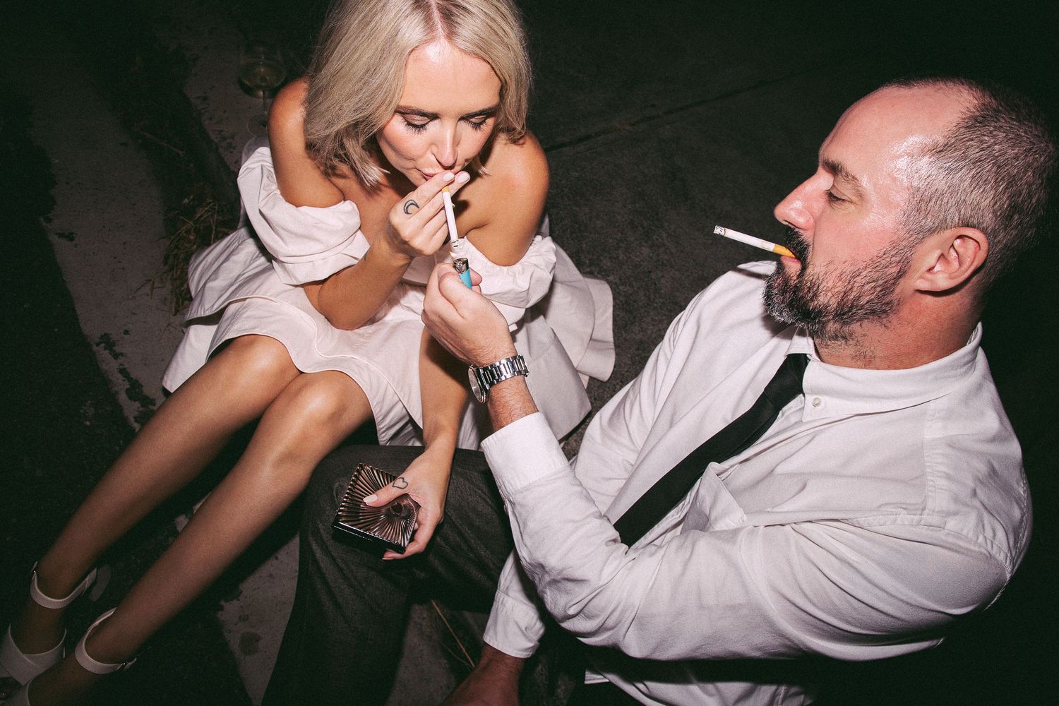 Groom lighting brides cigarette at dark wedding reception, both in formal white attire with drinks in hand.