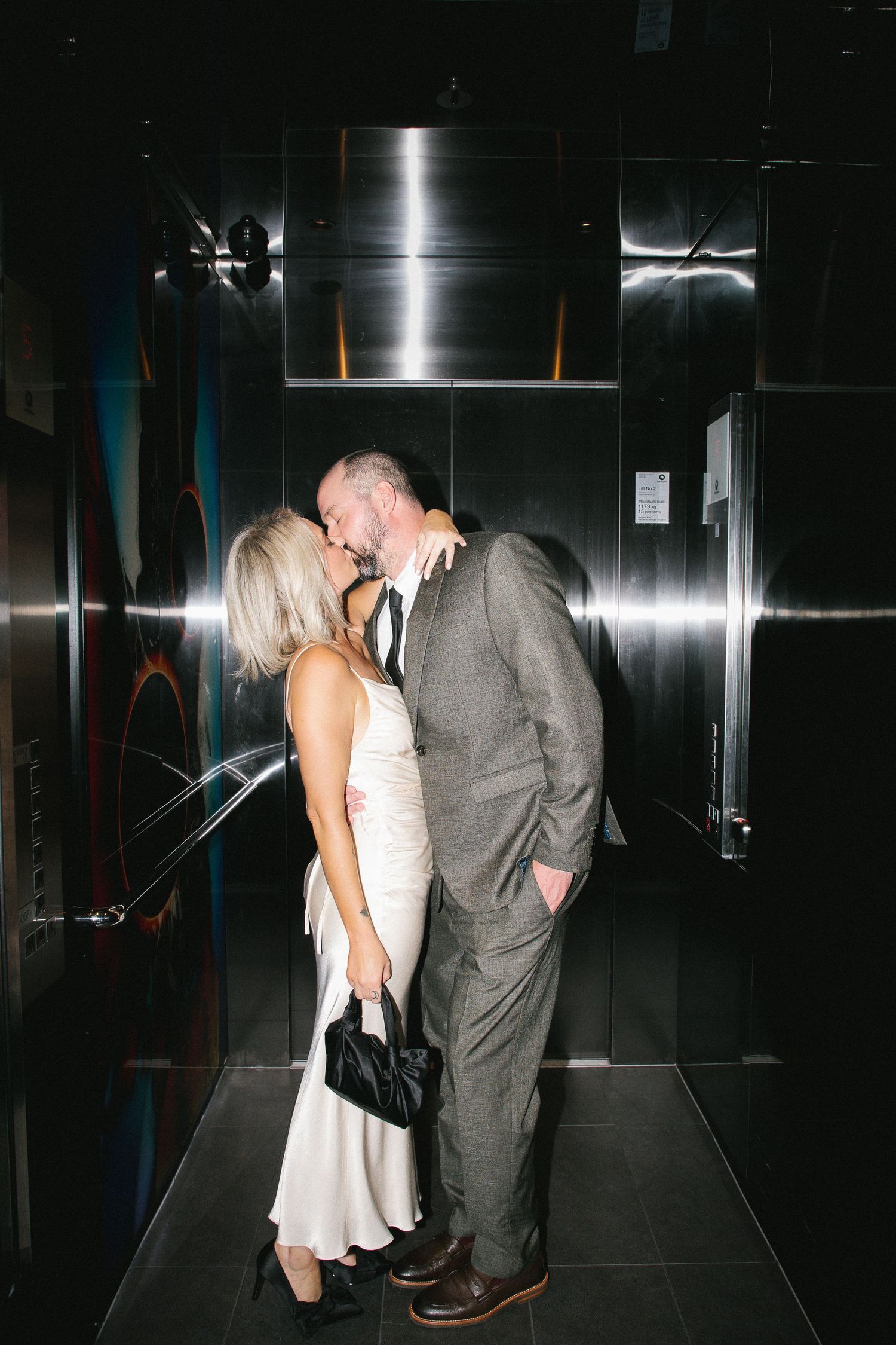 Newlywed couple sharing romantic kiss in elevator, bride in white silk dress holding black clutch purse.