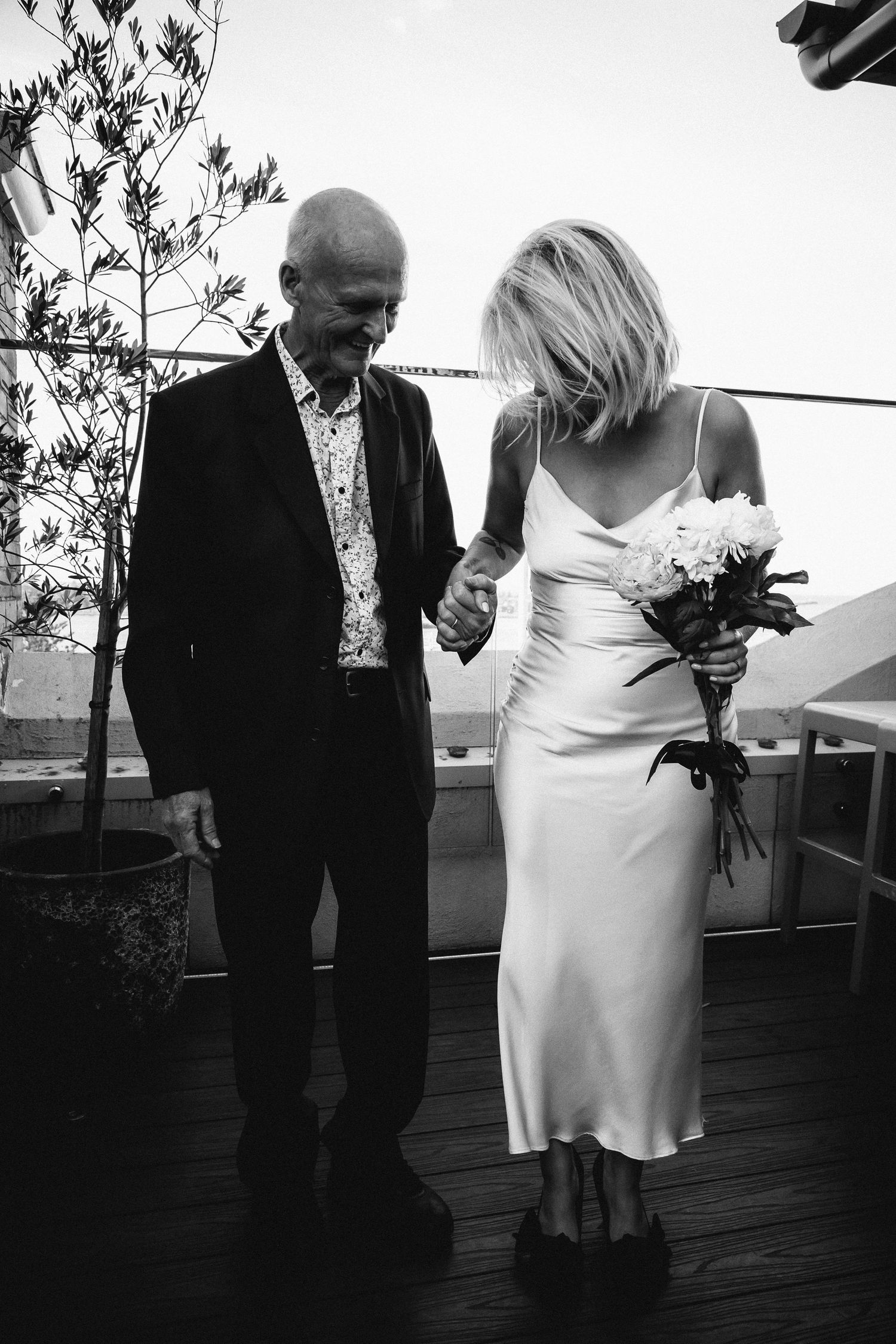 Black and white photo of bride in satin slip dress holding bouquet walking with elderly man on rooftop terrace.