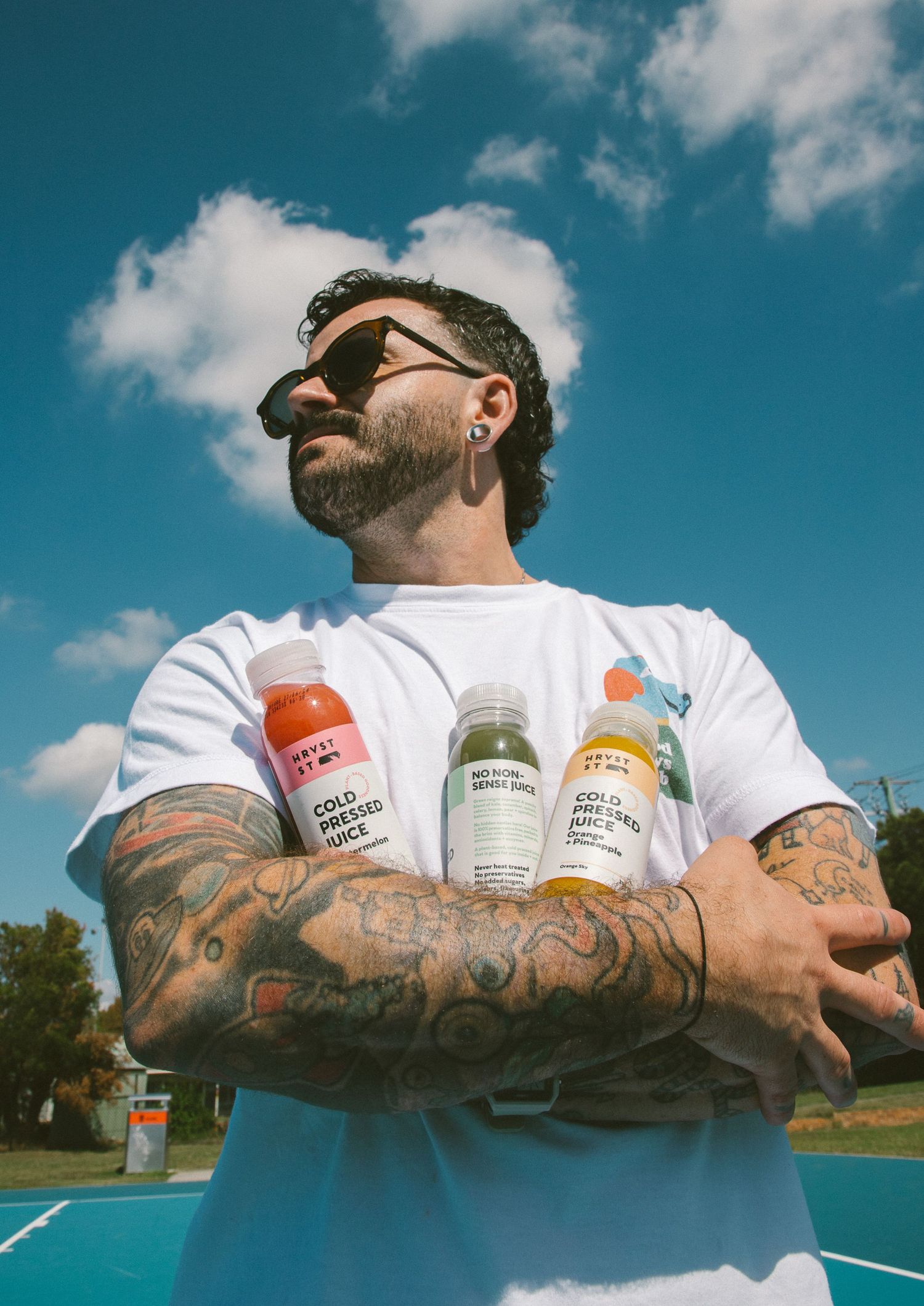 Tattooed man in white tee holding three cold pressed juice bottles with arms crossed under blue sky.