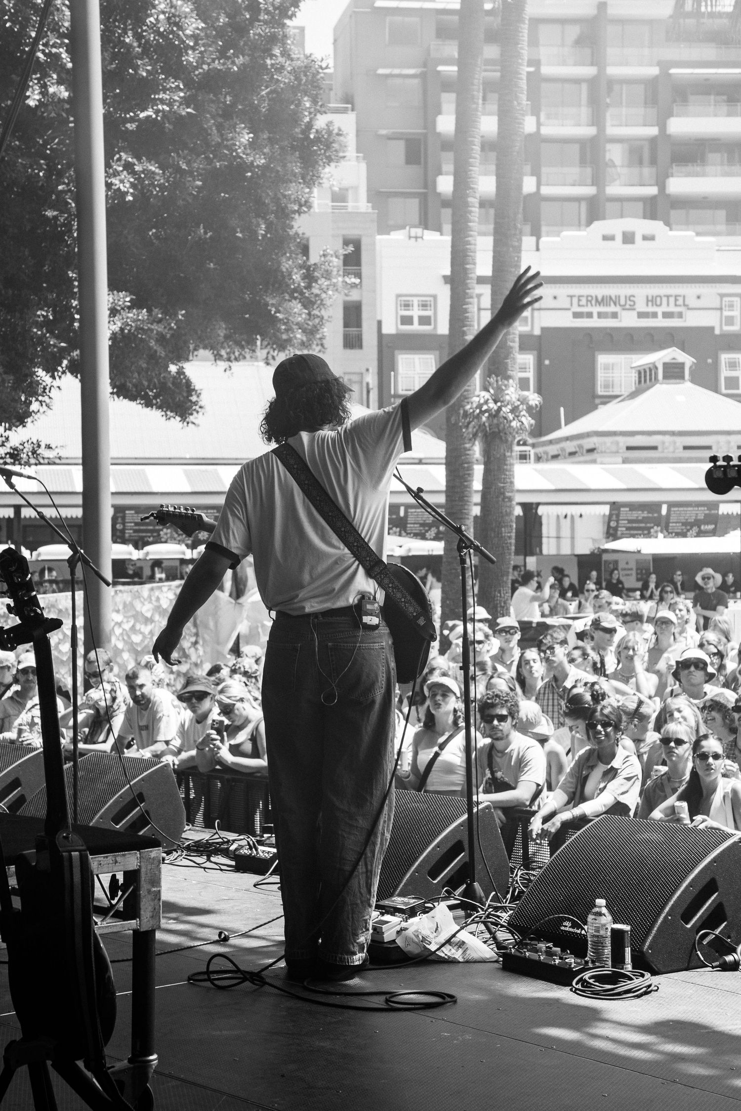 Black and white shot of performer raising hand to large outdoor festival crowd from behind on stage.