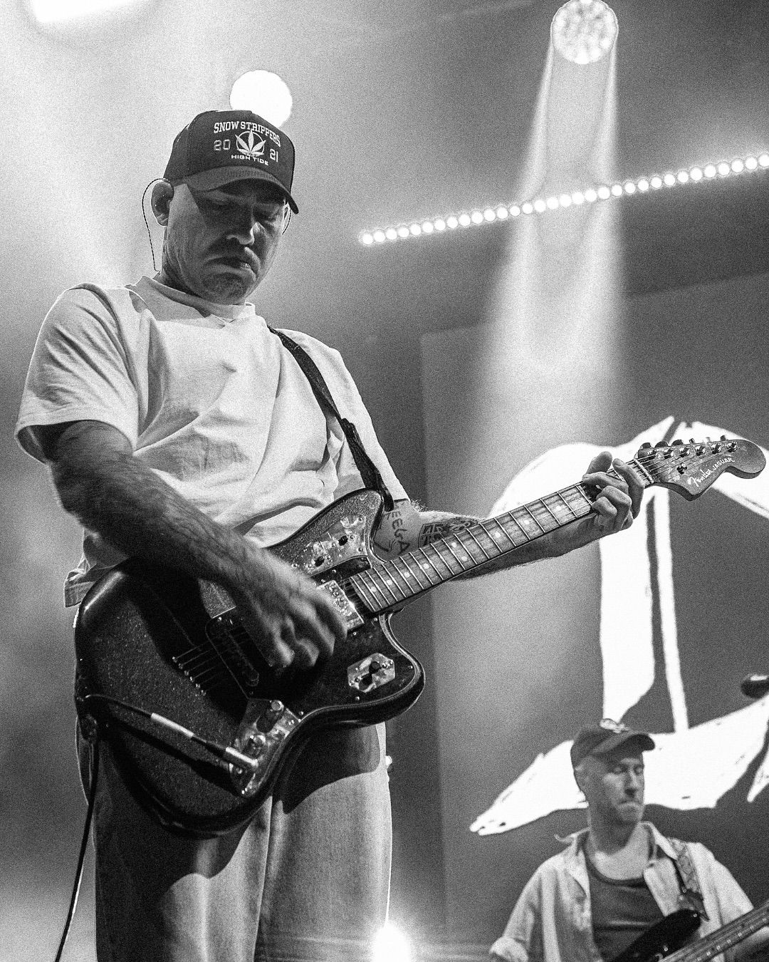 Black and white concert photo of DMAs guitarist in cap playing Fender guitar under bright stage lights.
