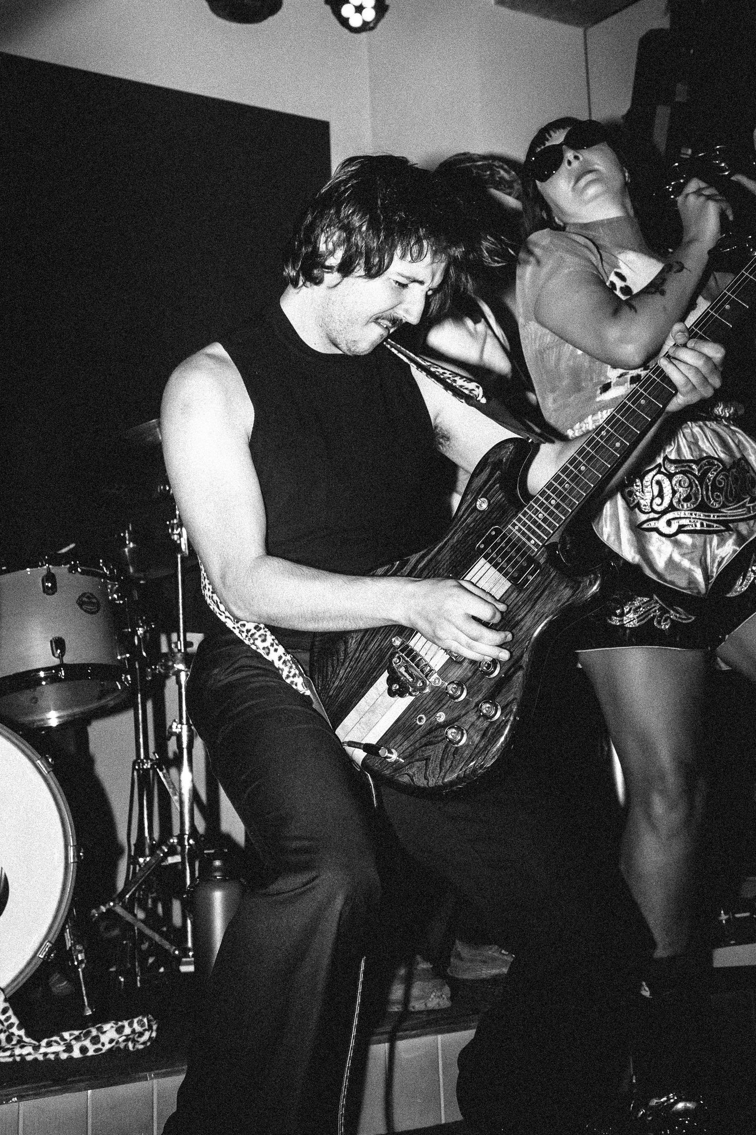 Black and white photo of rock musicians performing on stage, guitarist playing electric guitar with drummer in background.