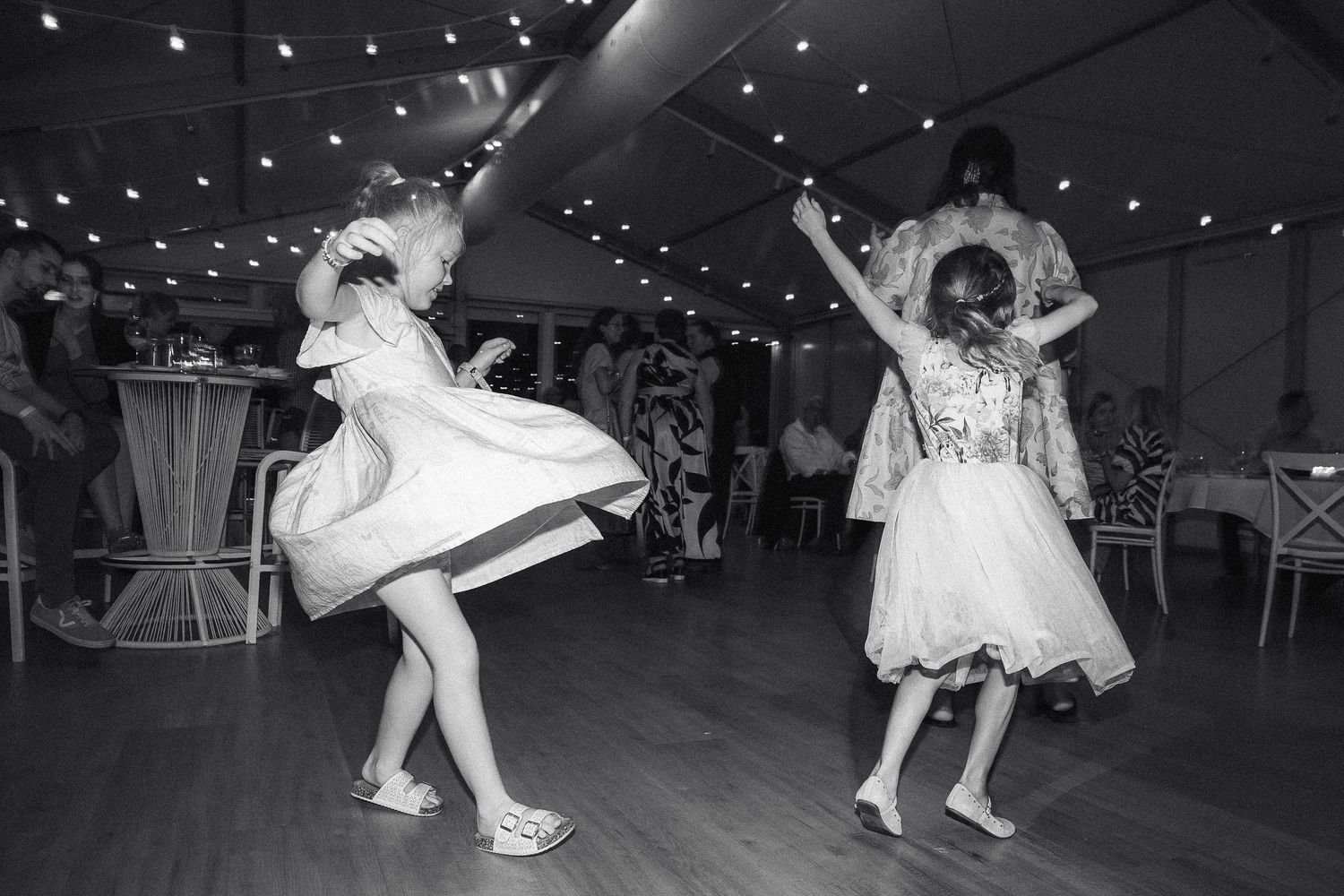 Black and white photo of two young girls in dresses dancing joyfully at wedding reception under string lights.