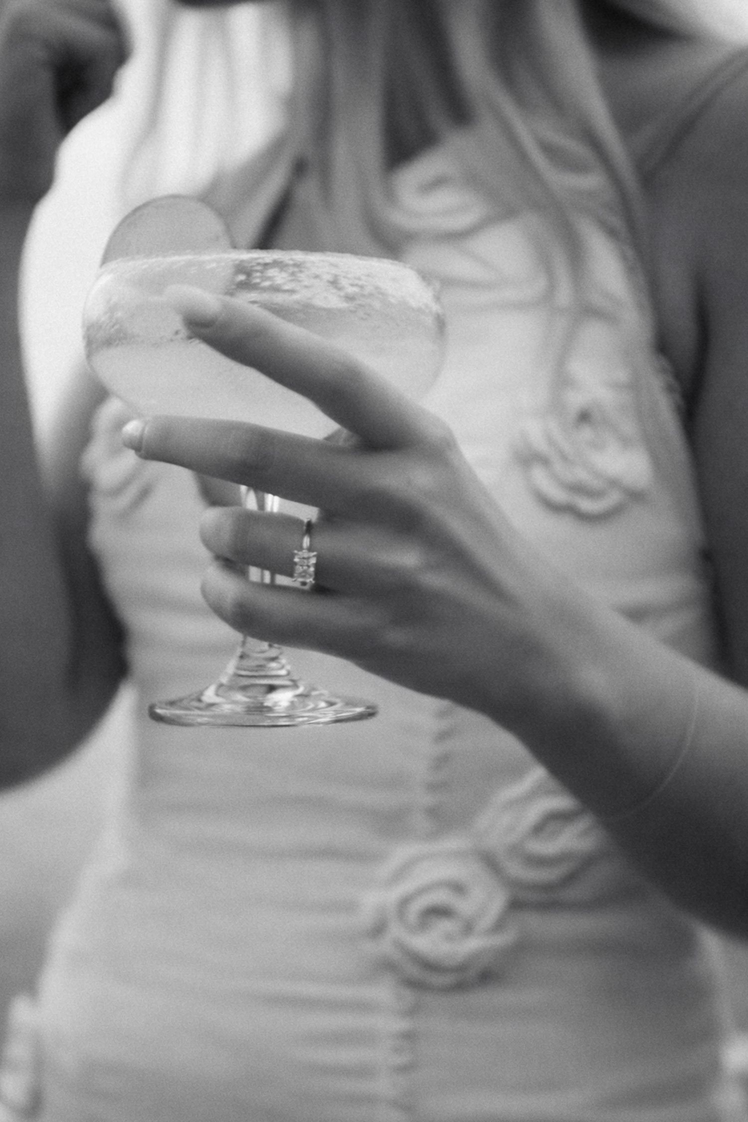 Black and white close-up of woman in floral dress holding champagne coupe glass with diamond ring visible.