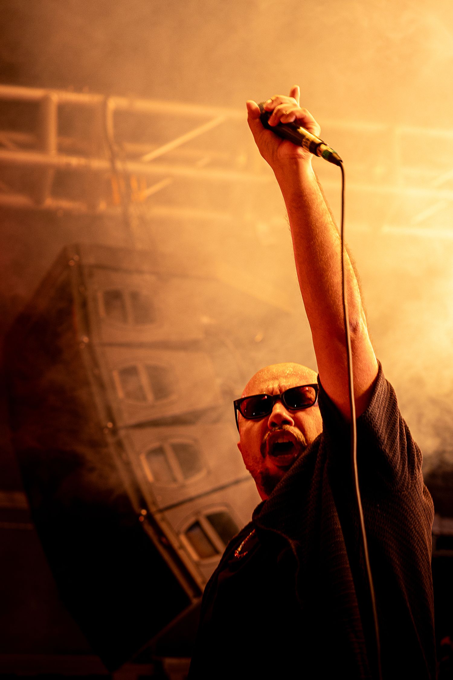 Bald performer raises microphone triumphantly on smoky stage wearing sunglasses during live concert.