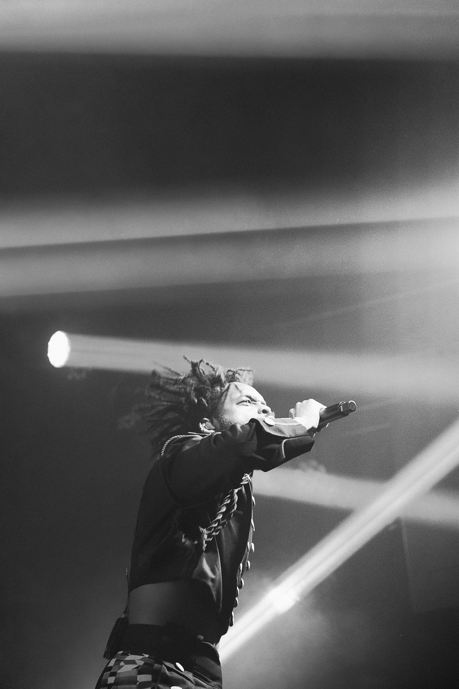 Black and white photo of a performer with dreadlocks singing into a microphone on stage with dramatic light beams.