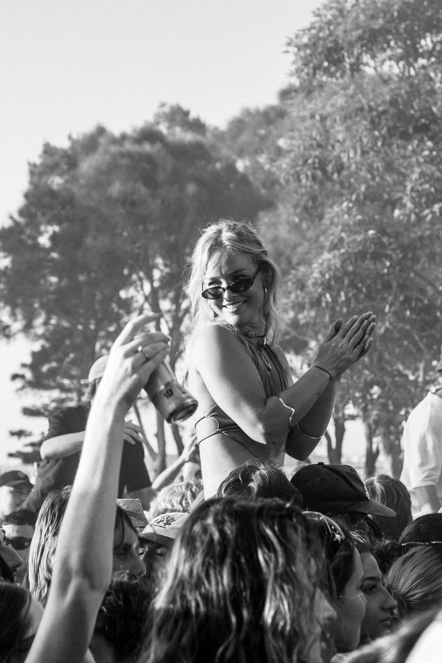 Black and white photo of smiling blonde woman in bikini top sitting on shoulders in outdoor Earp  festival crowd.