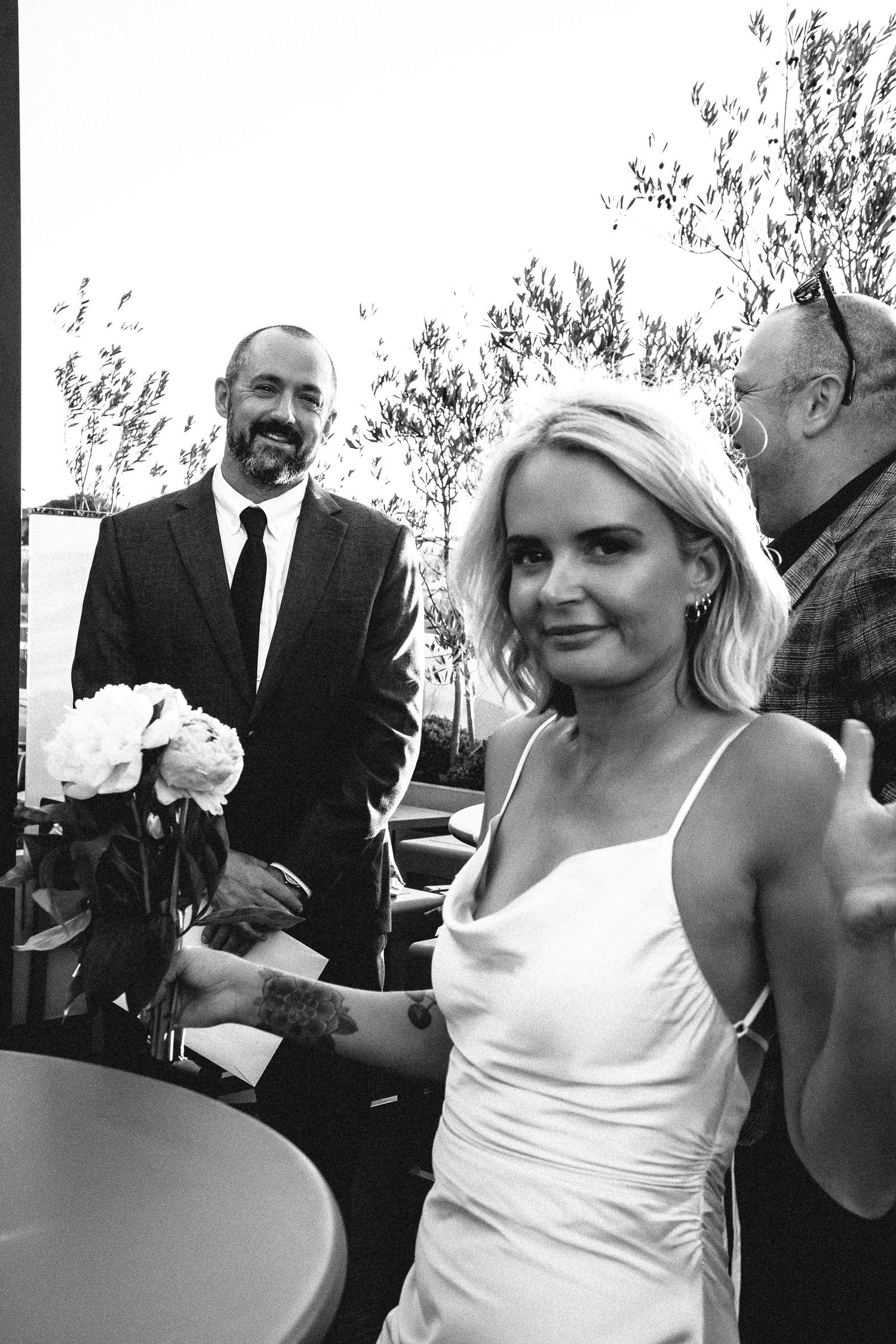Black and white candid of smiling groom in suit with bride holding white peonies at outdoor wedding reception.
