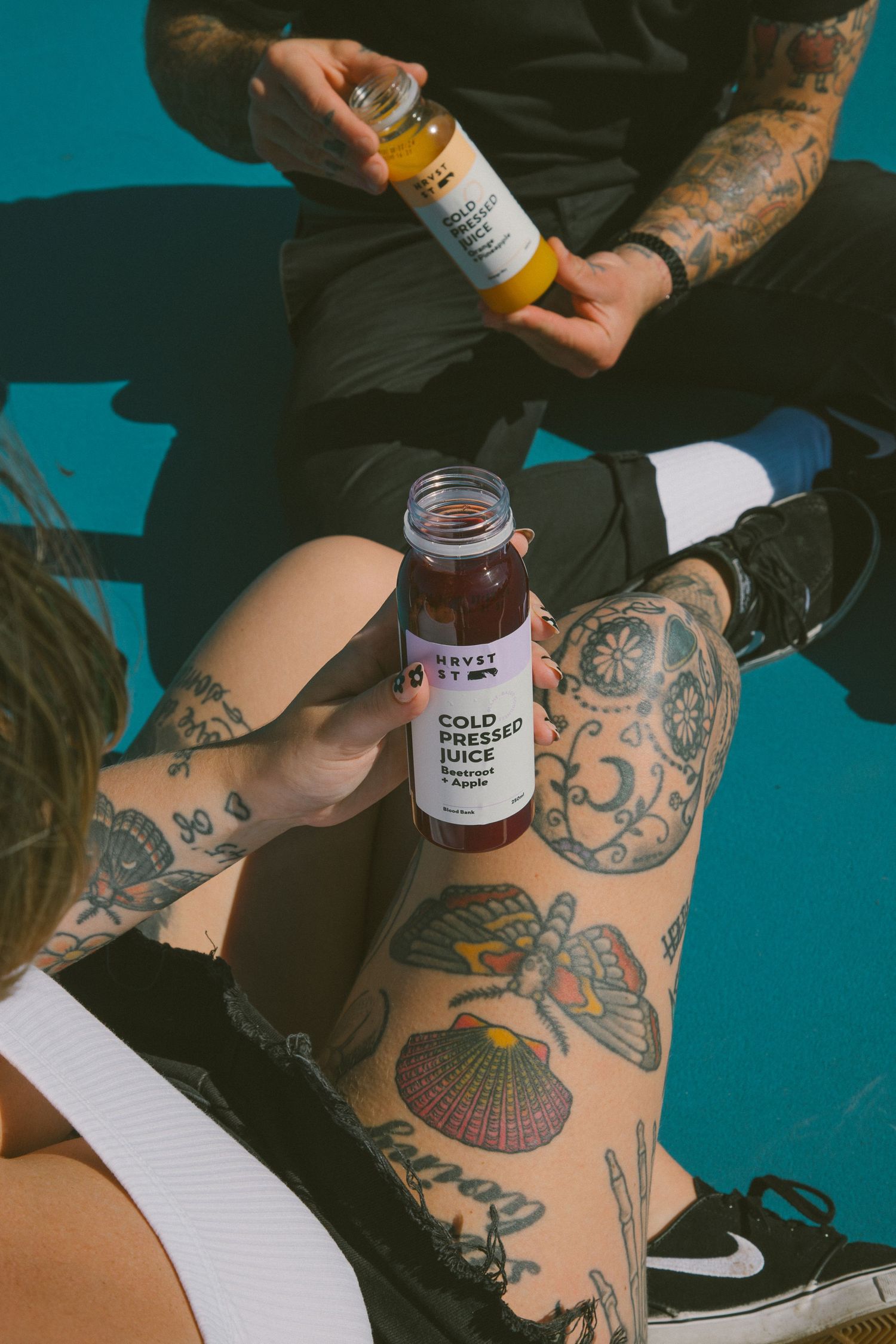Two tattooed people sitting on a tennis court holding HRVST ST Cold Pressed Juice bottles in beetroot and apple flavors.