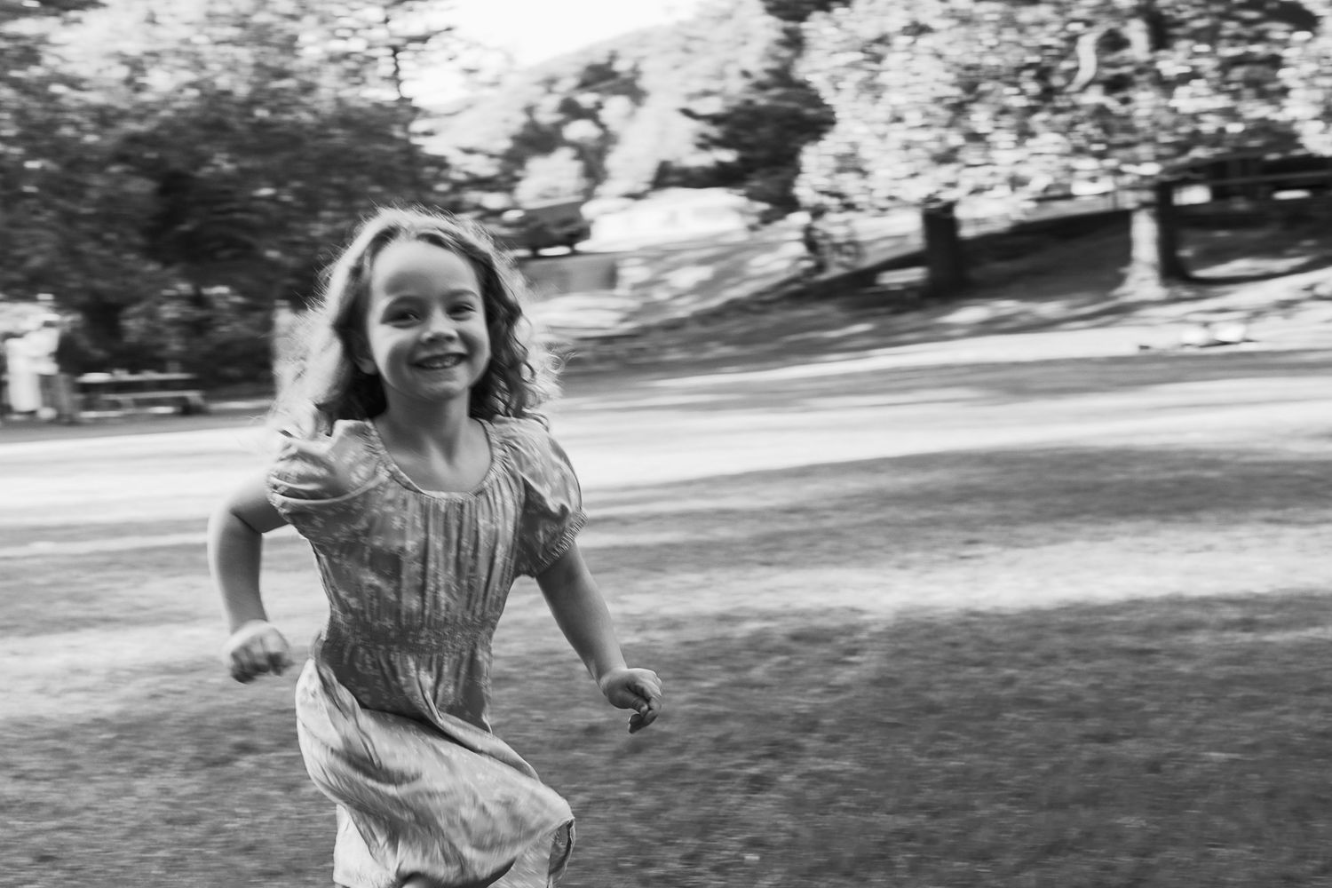 A joyful young girl in a dress runs outdoors in a black and white photo, smiling with arms outstretched.