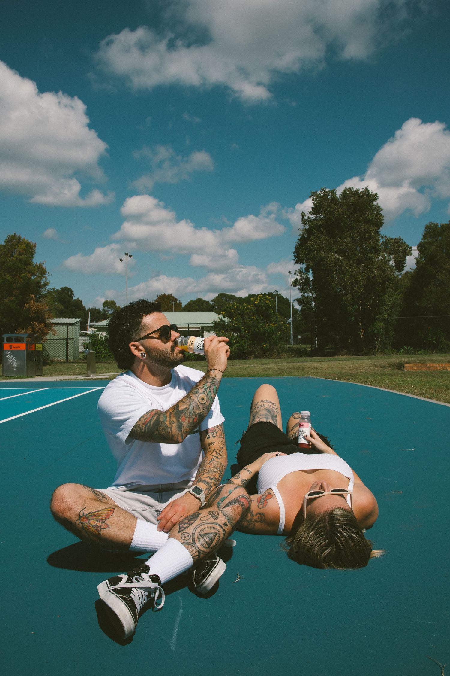 Tattooed couple sitting on blue basketball court drinking cold pressed juices on a sunny day.