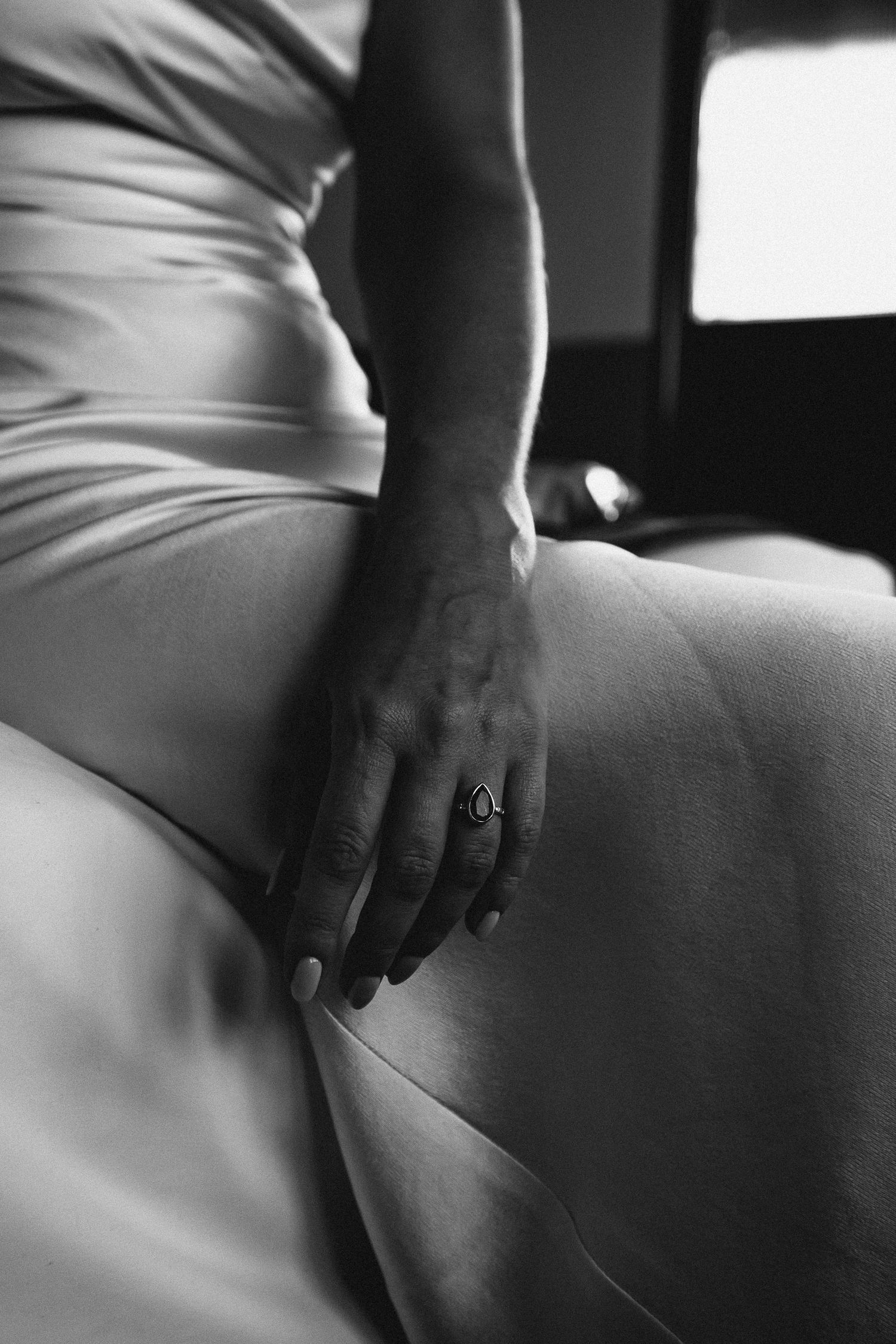 Close-up black and white photo of a hand with ring resting on white satin dress.