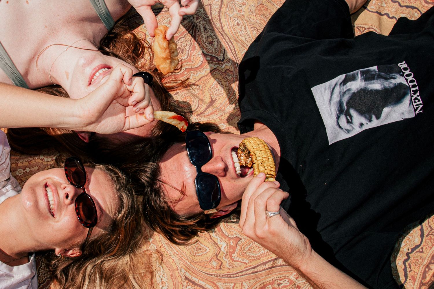 Three friends lying on a blanket outdoors, laughing and eating corn cobs while wearing sunglasses at Merewether Beach.