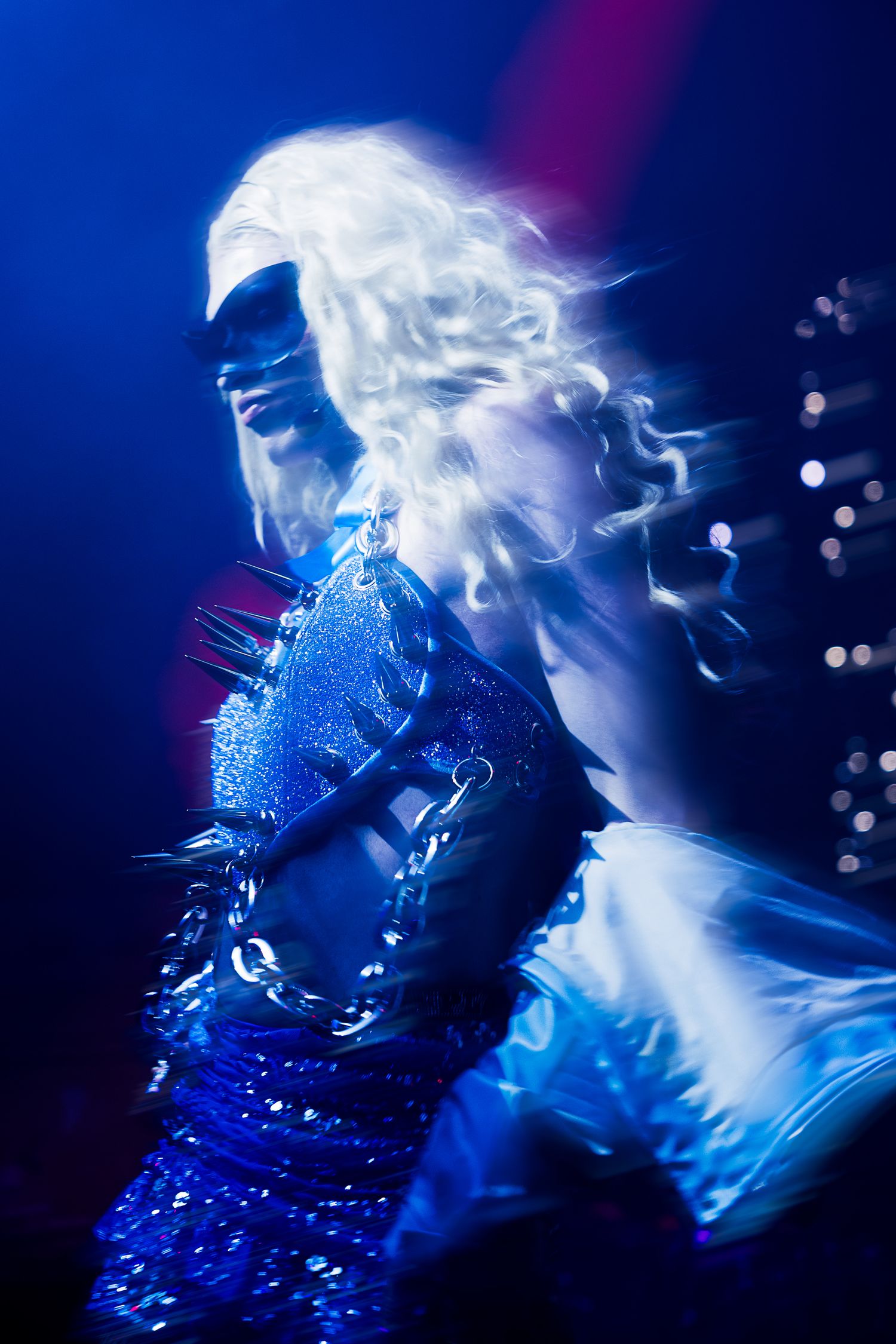 A performer in a blue sequined costume and white wig dances under dramatic blue stage lighting.