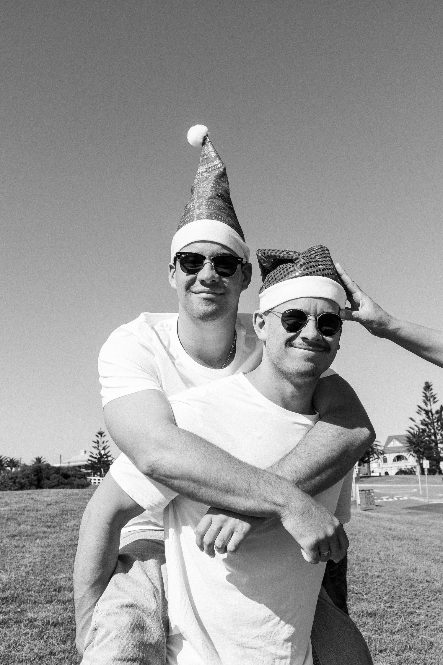 Two men wearing Santa hats and sunglasses pose for a piggyback photo outdoors in black and white.