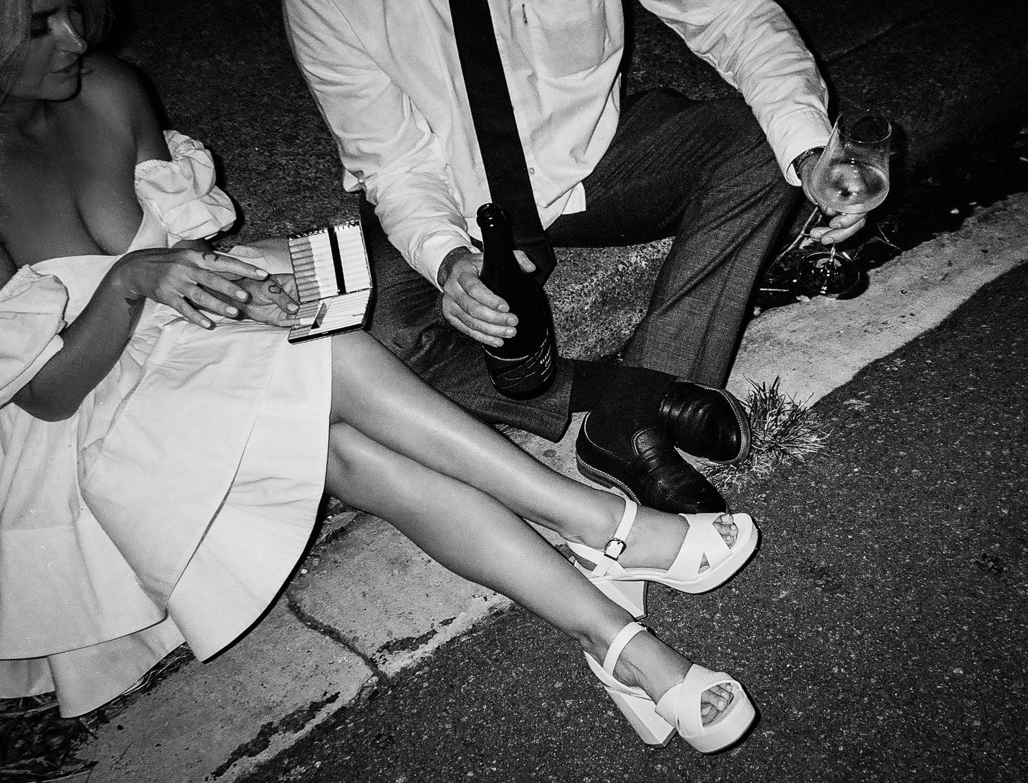 Black and white photo of couple sitting on curb in formal attire, holding drinks at outdoor event.