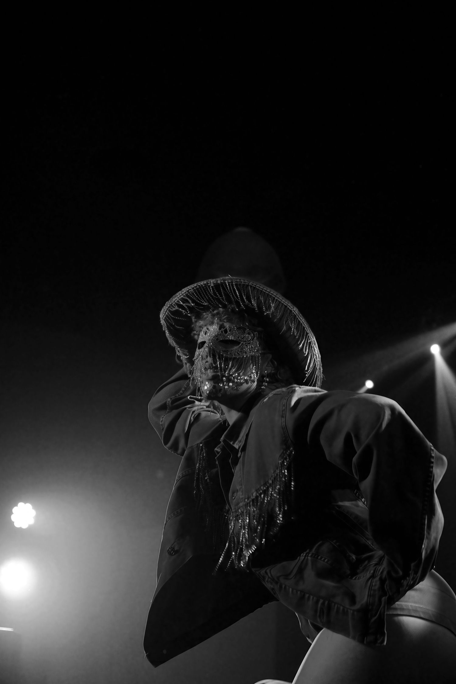 A masked performer in a wide-brimmed hat stands on stage under dramatic black and white lighting at The Station Hotel for Poof Doof.