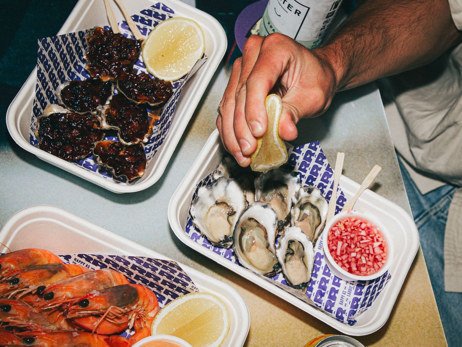Hands squeezing lemon over fresh oysters on a tray, with prawns and caviar on a seafood platter at Larrie's Takeaway Merewether.
