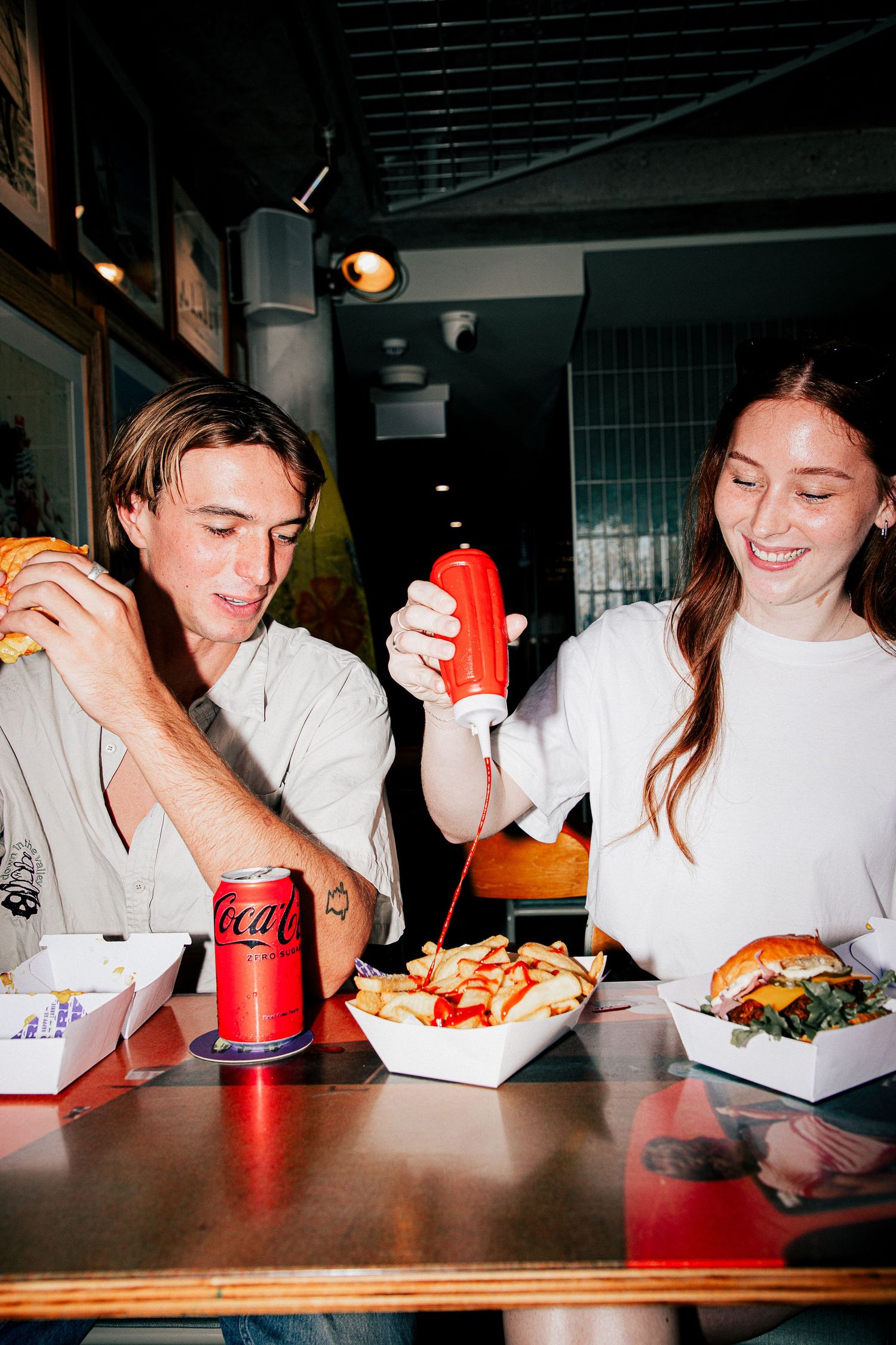Young people at Larrie's Takeaway Merewether with woman squeezing ketchup on fries beside burgers and a Coca-Cola can.