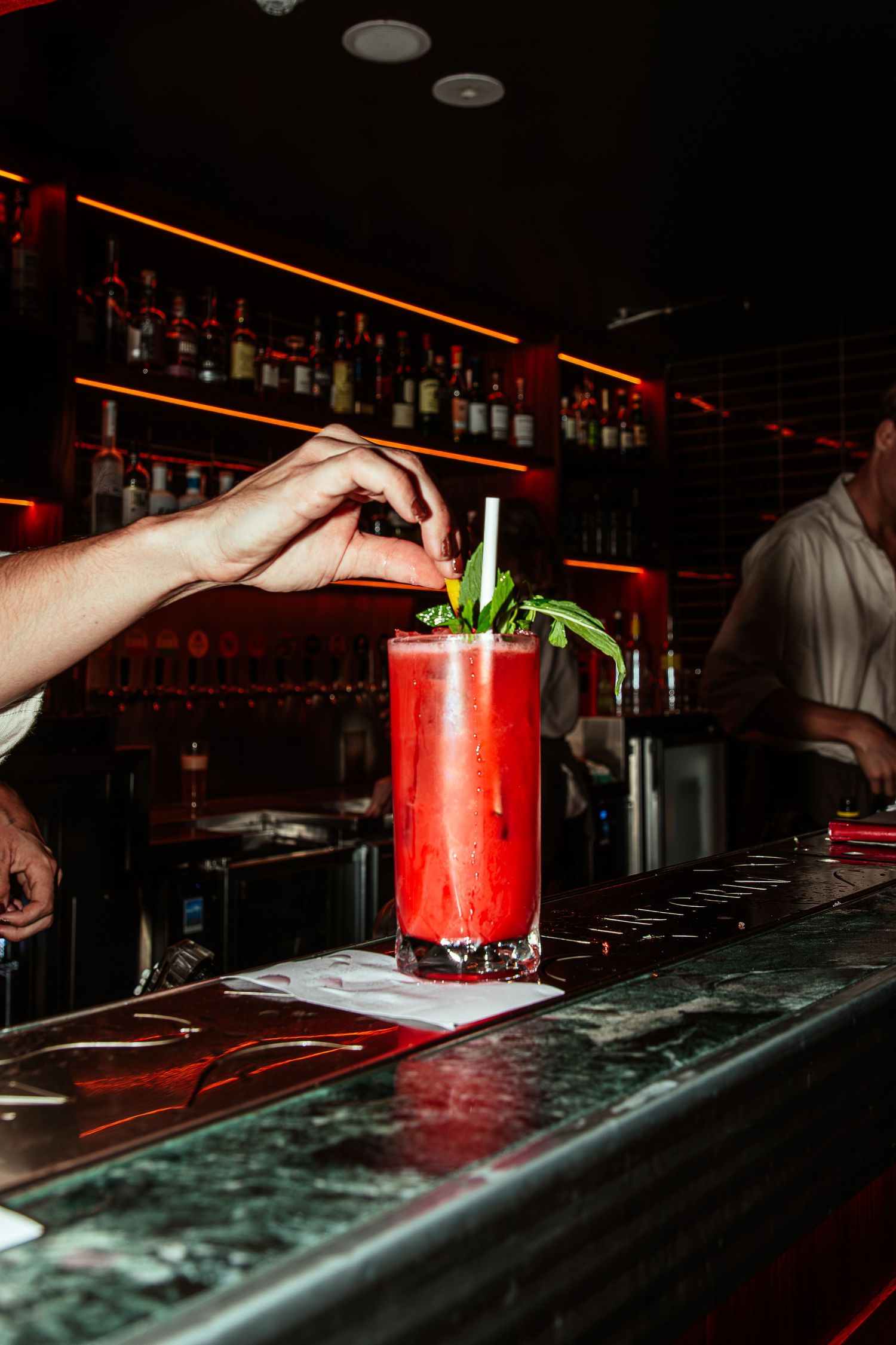 Bartender garnishing a red cocktail with fresh herbs at a dimly lit bar.