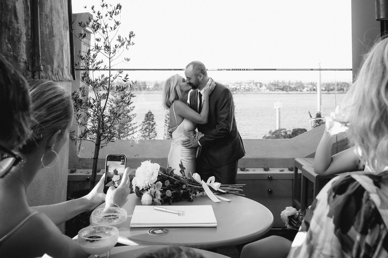Black and white image of guests watching newlyweds kiss over wedding registry at intimate rooftop ceremony.
