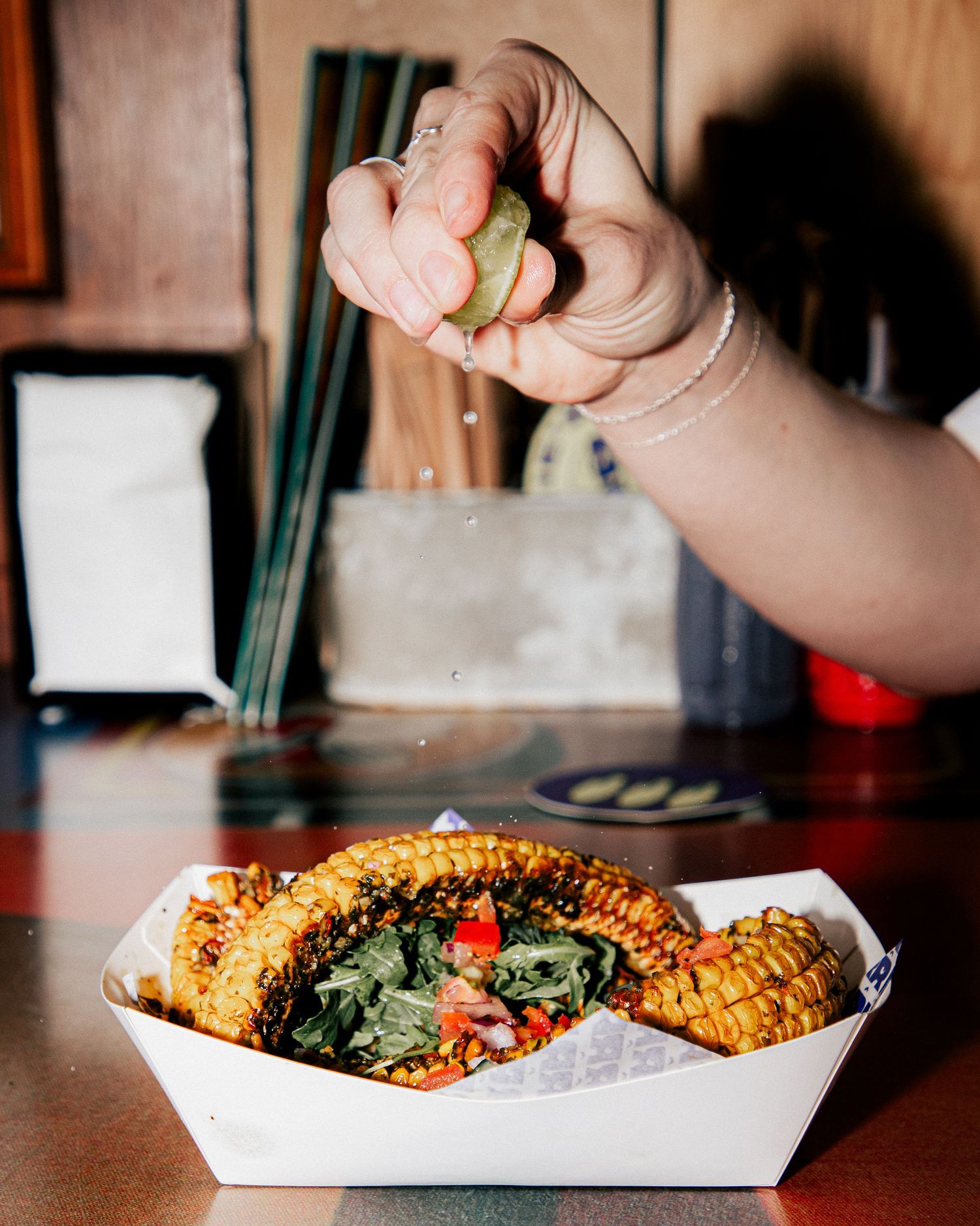 Hand squeezing fresh lime juice over a paper tray of grilled corn cobs with vegetables on a counter at Larrie's Takeaway Merewether.