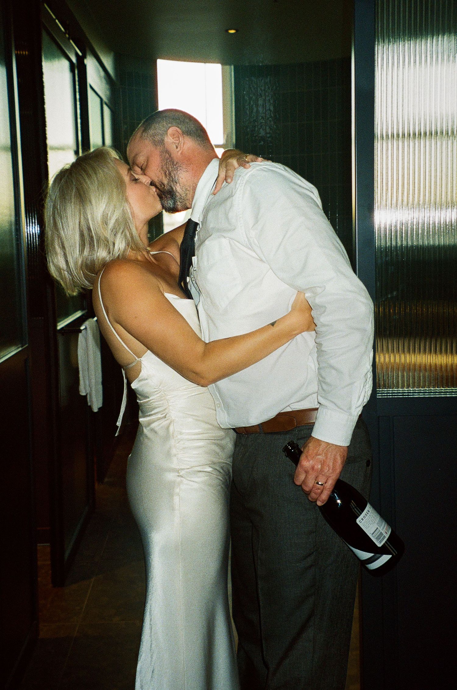 Bride in white satin gown and groom kissing in hallway, groom holding wine bottle at wedding reception.