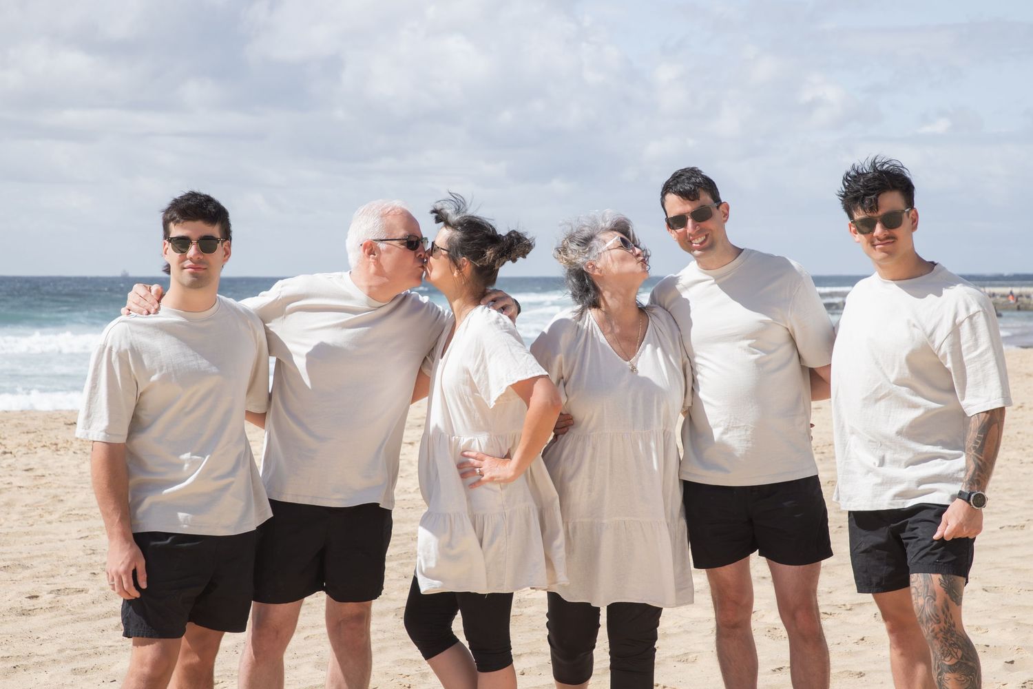 Group of six people in matching white shirts and black shorts posing together on a sunny beach.