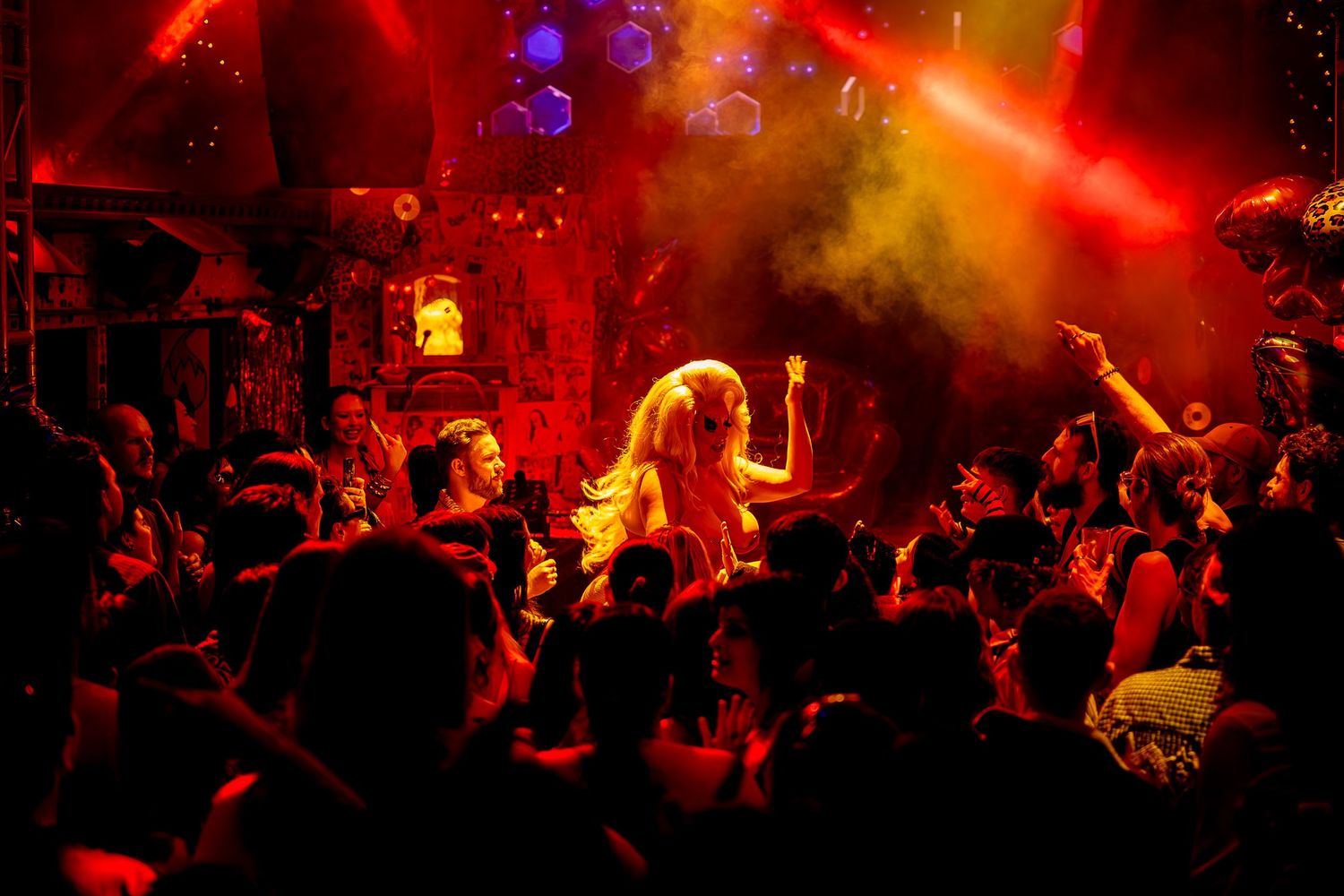 Crowd dancing under red and yellow lights at a vibrant nightclub with a blonde performer on stage.