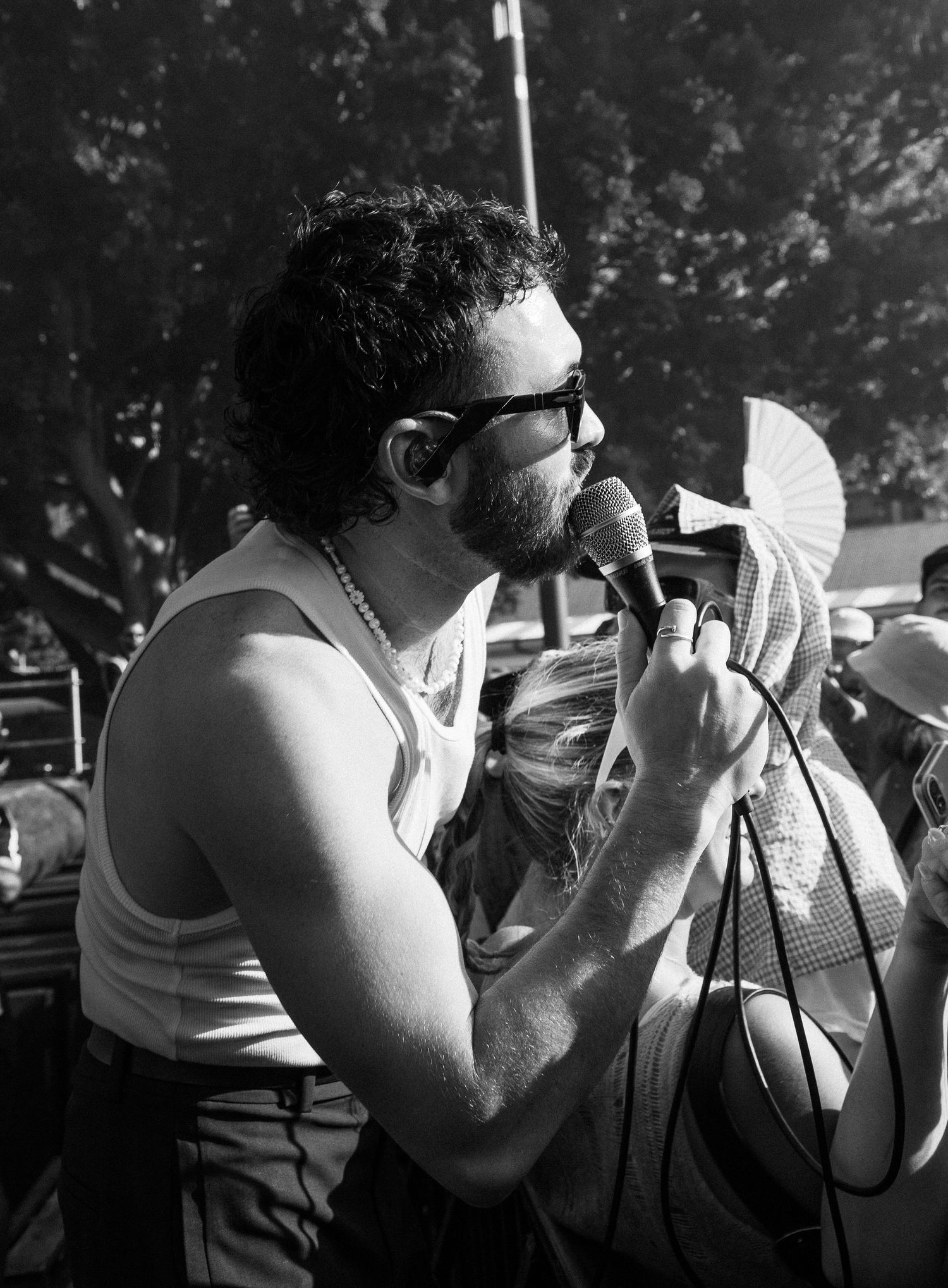 Black and white close-up of bearded male lead singer of San Cisco in tank top performing with microphone at outdoor event.