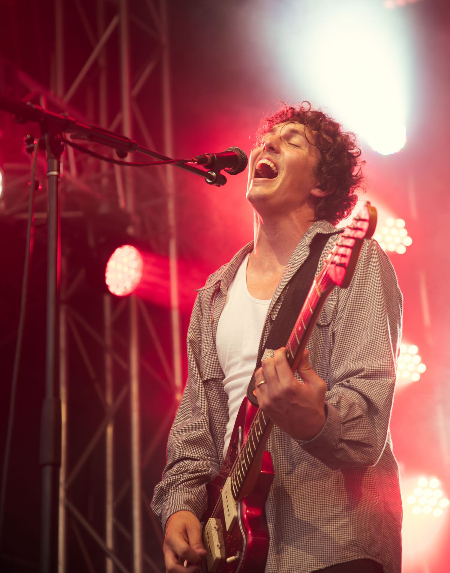 Curly-haired male guitarist of Hockey Dad band singing passionately on stage with red electric guitar under dramatic red stage lighting.