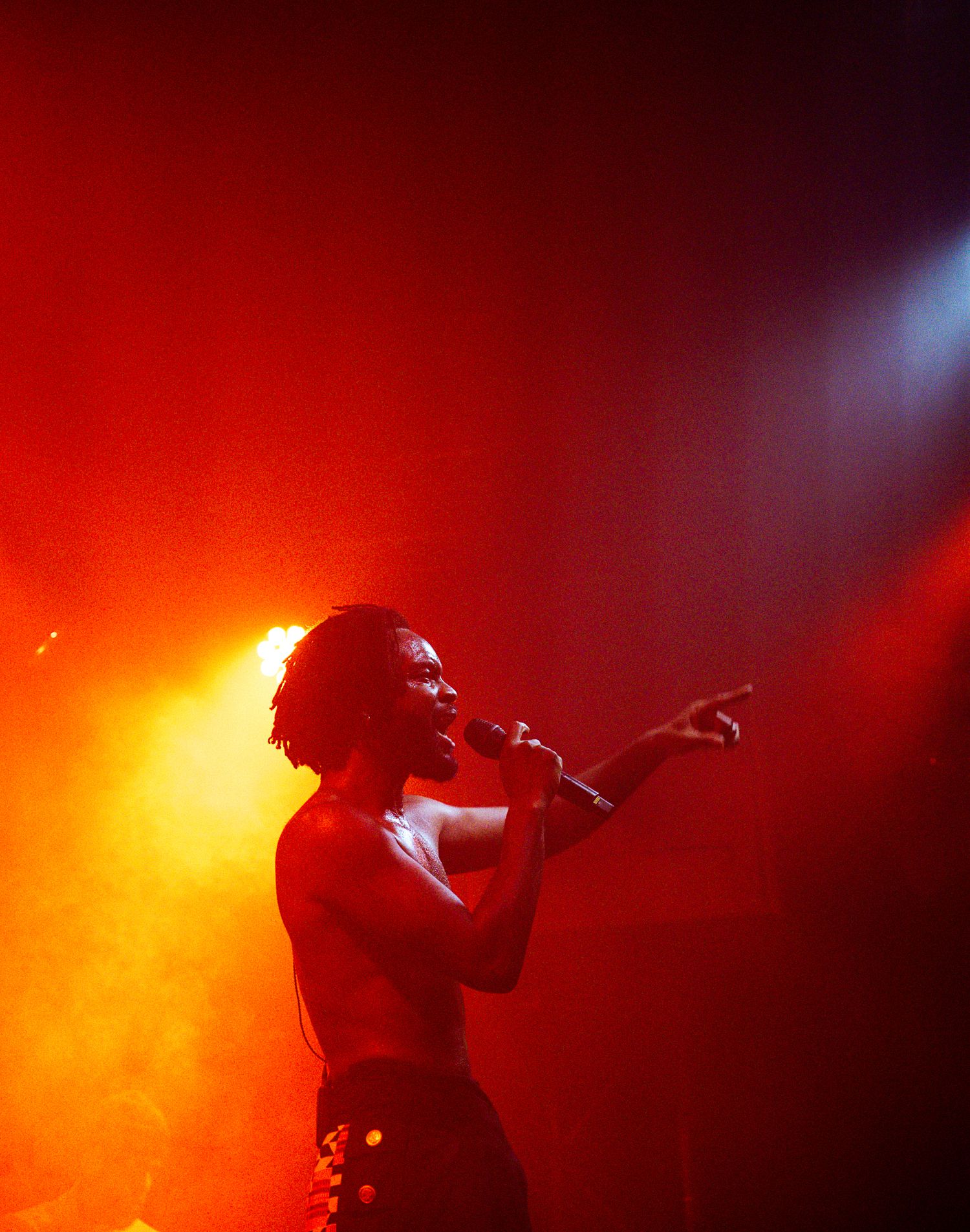 A shirtless performer sings into a microphone on a dramatic red-lit stage, pointing outward during a live concert.