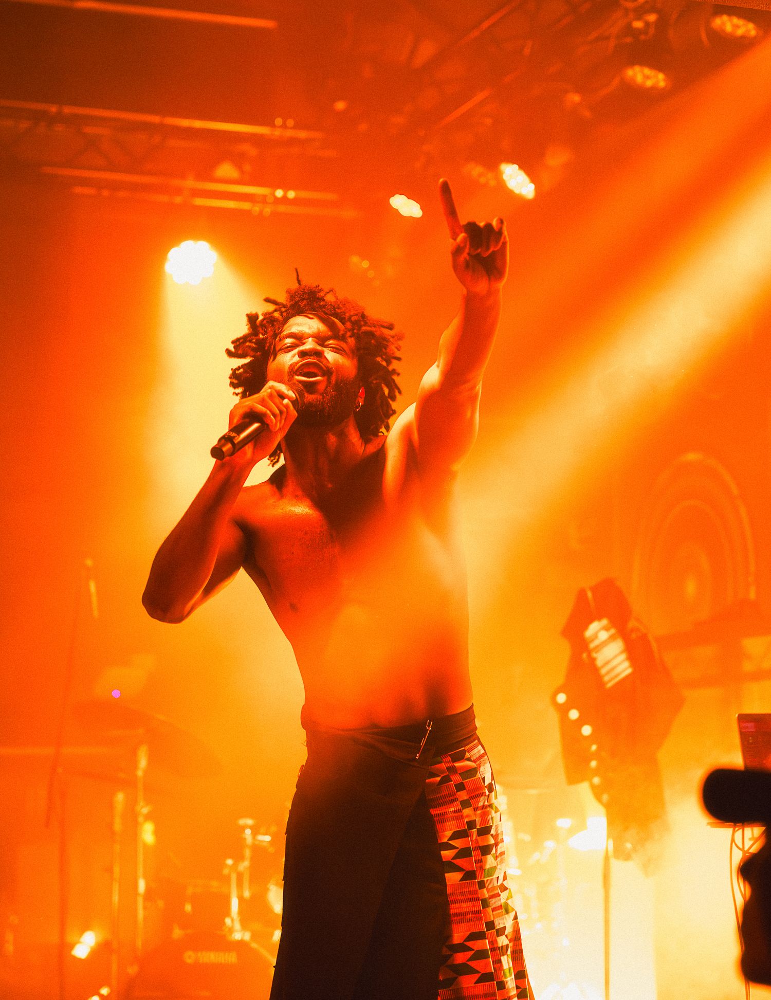 Shirtless male singer performing on stage with microphone, raising finger under dramatic orange stage lighting.