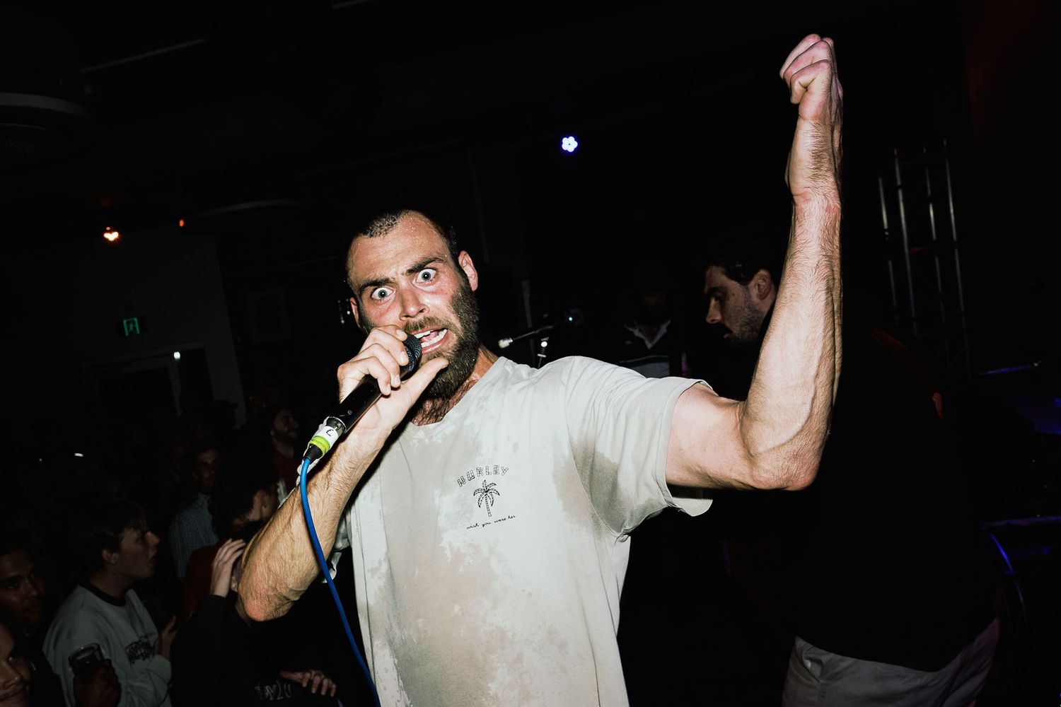 Male singer performing on stage with microphone, fist raised, wearing white t-shirt in dark nightclub venue.
