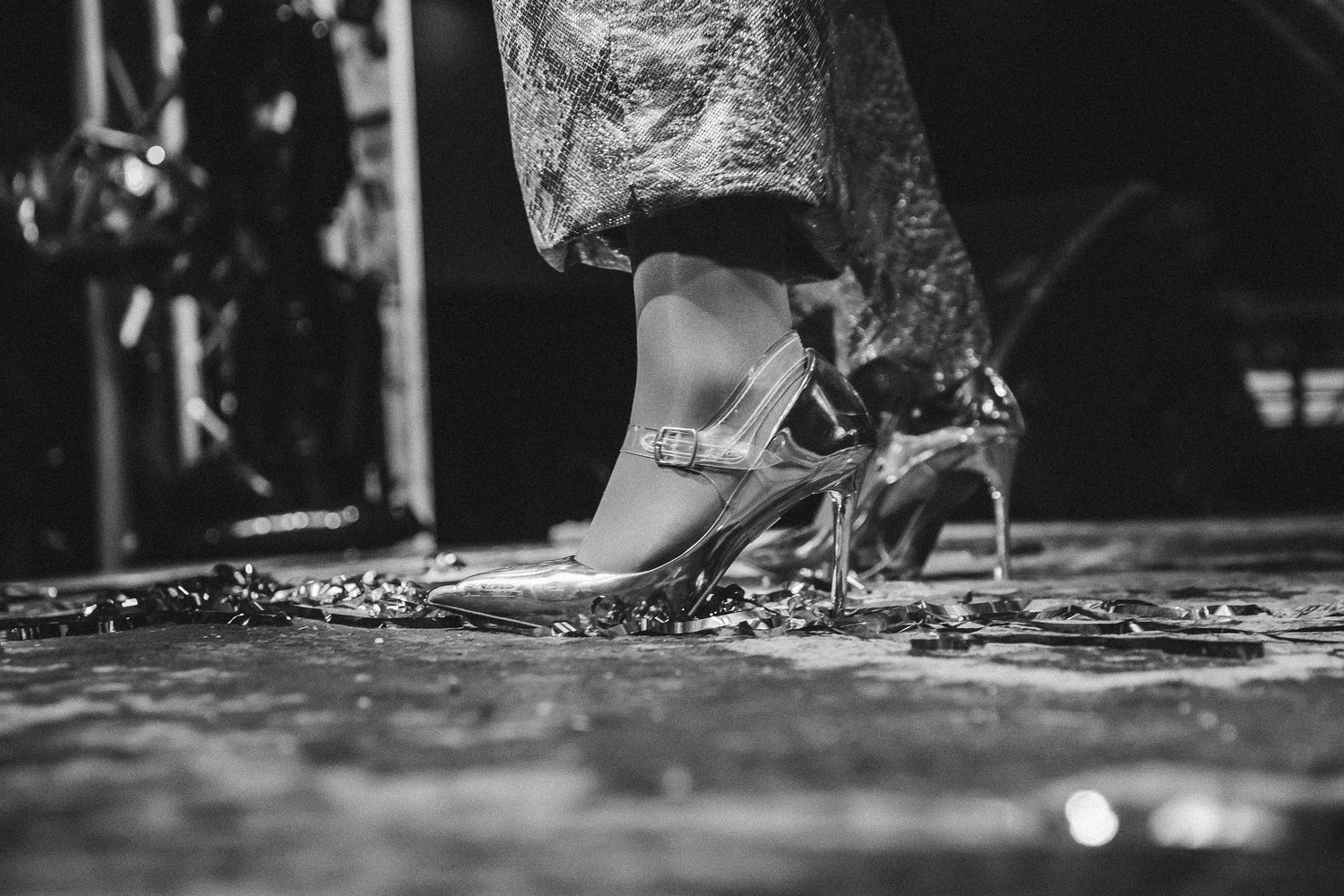 Black and white close-up of womans feet in clear heels on glitter-covered floor.
