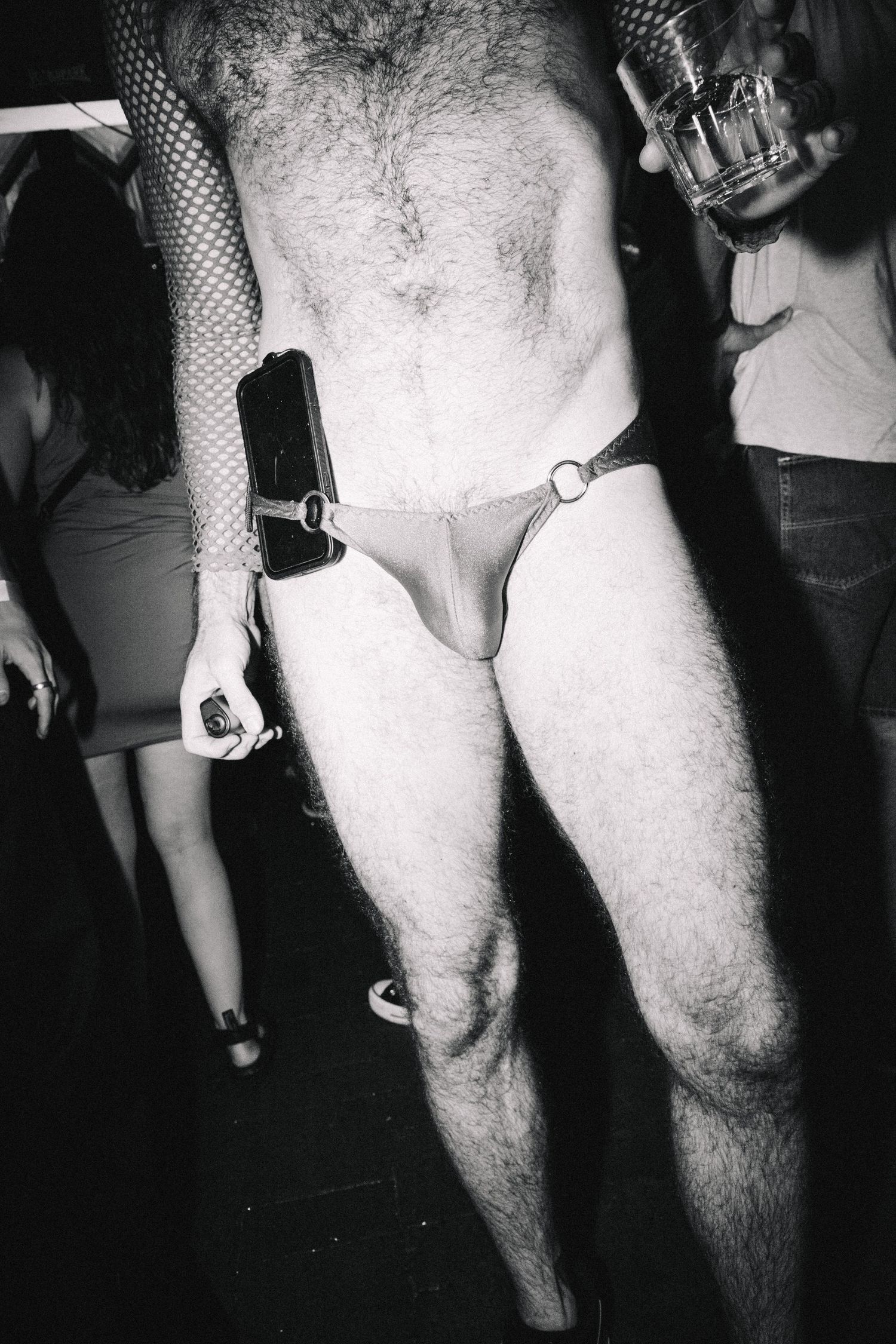Black and white close-up of a person wearing a minimal thong harness at a nightclub event at The Lass O'Gowrie Hotel.