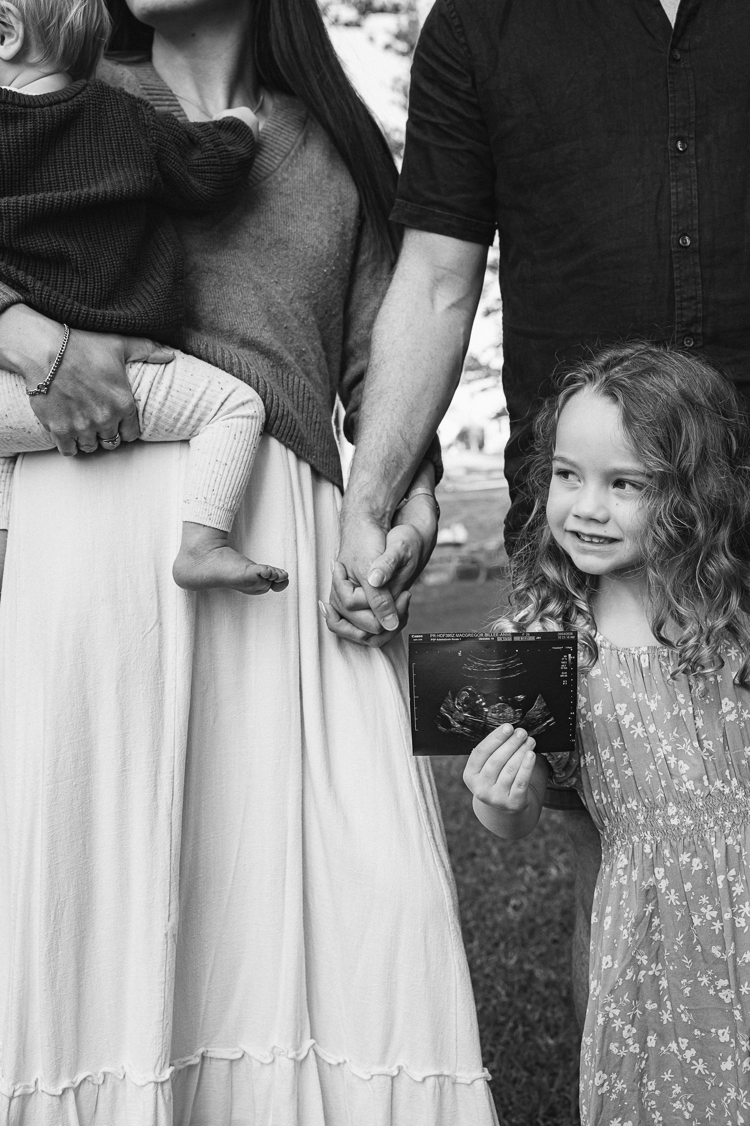 Black and white family photo with parents holding hands and curly-haired girl displaying ultrasound photo outdoors.