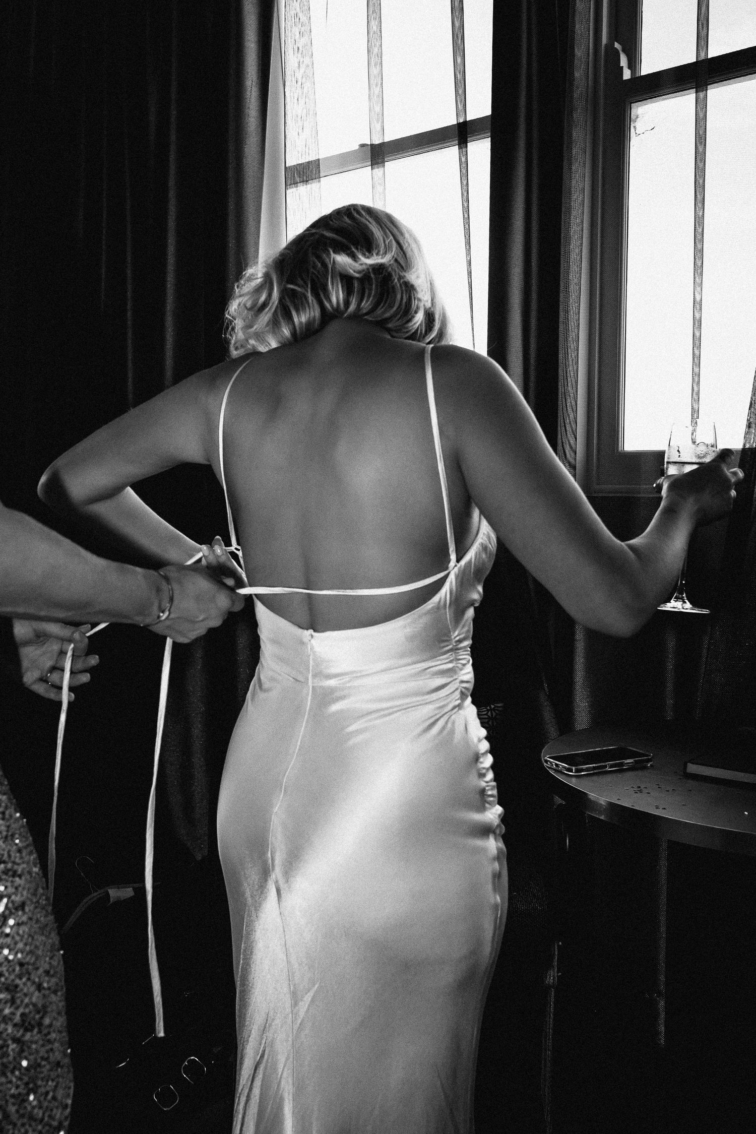 Black and white photo of bride in backless silk wedding dress being tied up before intimate ceremony.