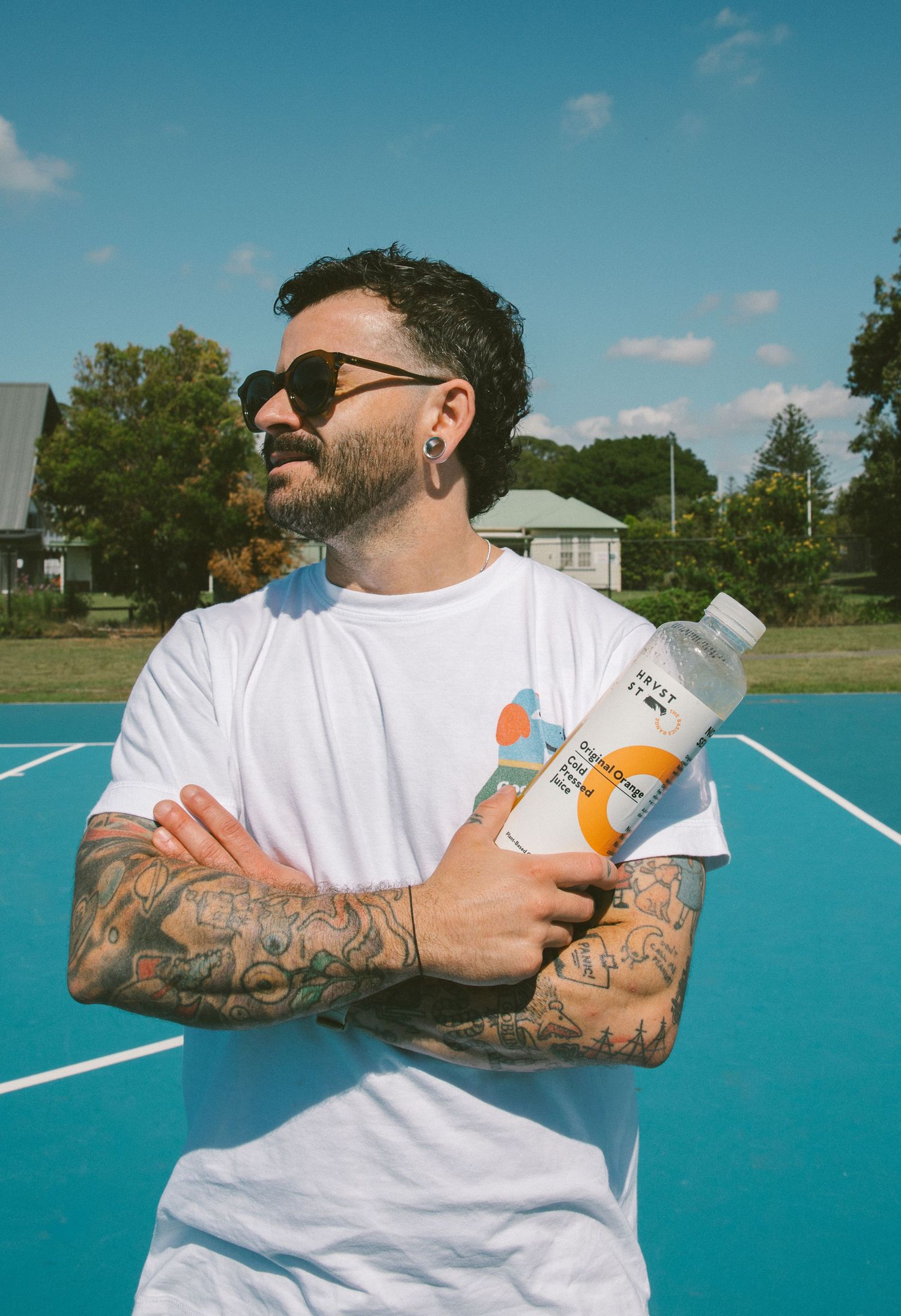 Tattooed man in white t-shirt holding cold pressed juice bottle with arms crossed on blue court.