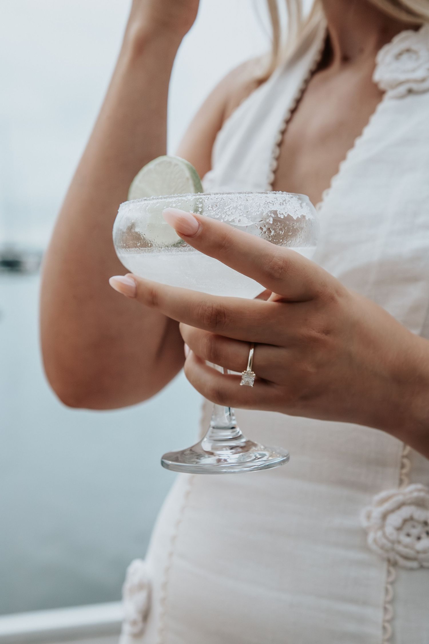 Bride in white crochet dress holding margarita glass with lime garnish near waterfront at wedding celebration.