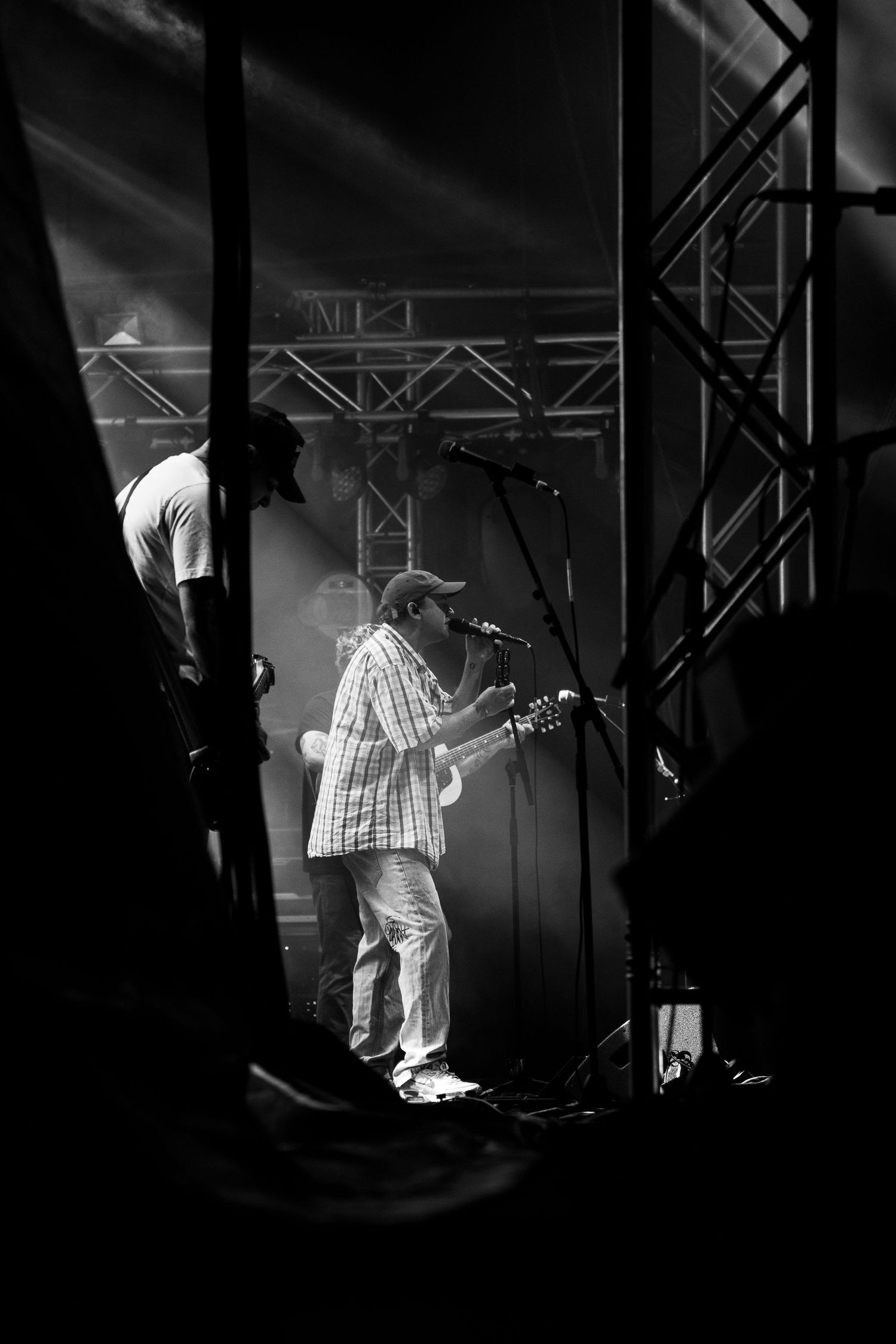 Black and white backstage view of lead singer of DMAs in plaid shirt performing under dramatic stage lighting and fog.