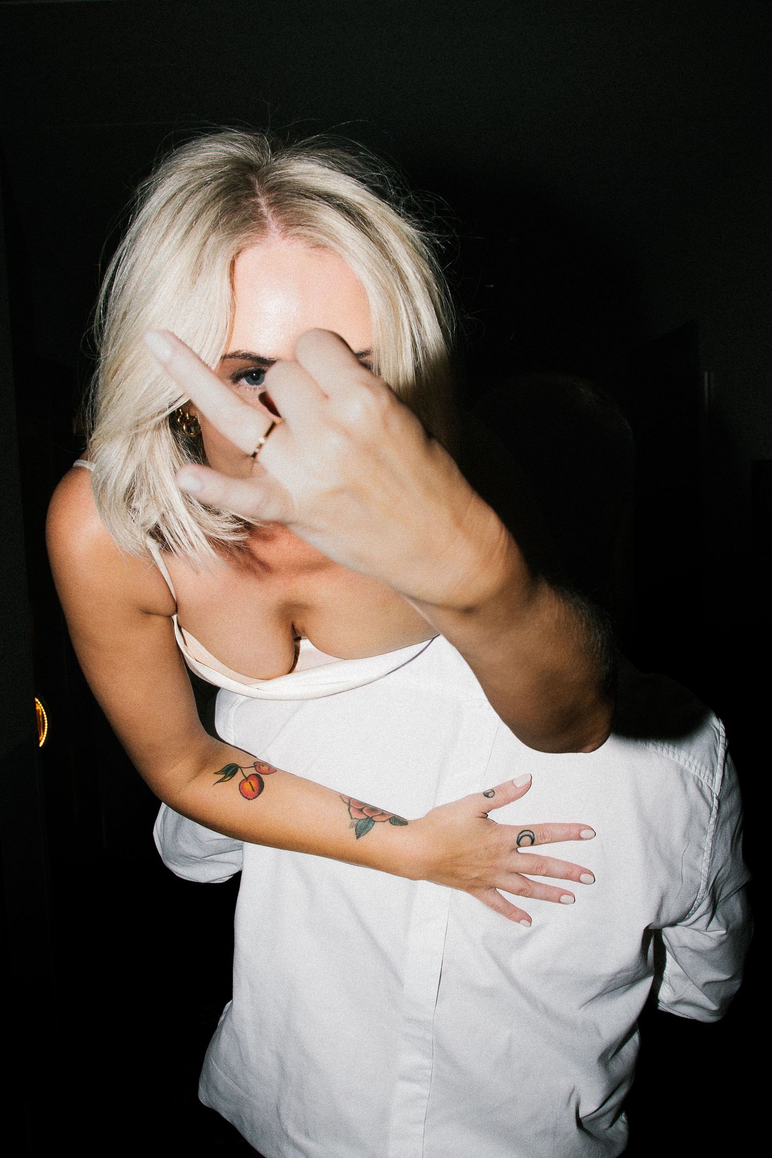 Bride in white dress with cherry tattoo embraces groom in white shirt, his hand playfully covering camera.