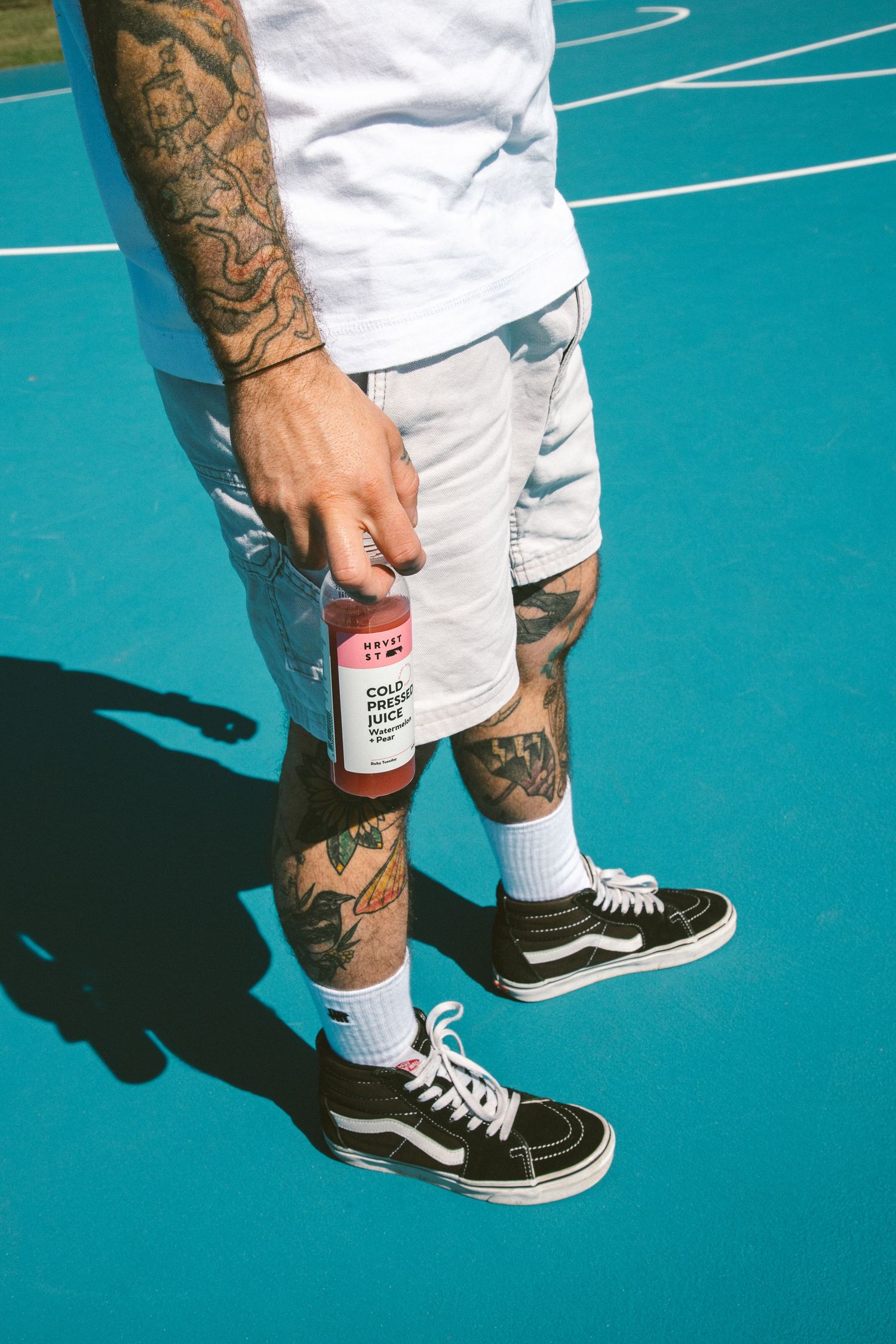 Tattooed legs in white shorts and Vans sneakers holding cold pressed juice on blue court.
