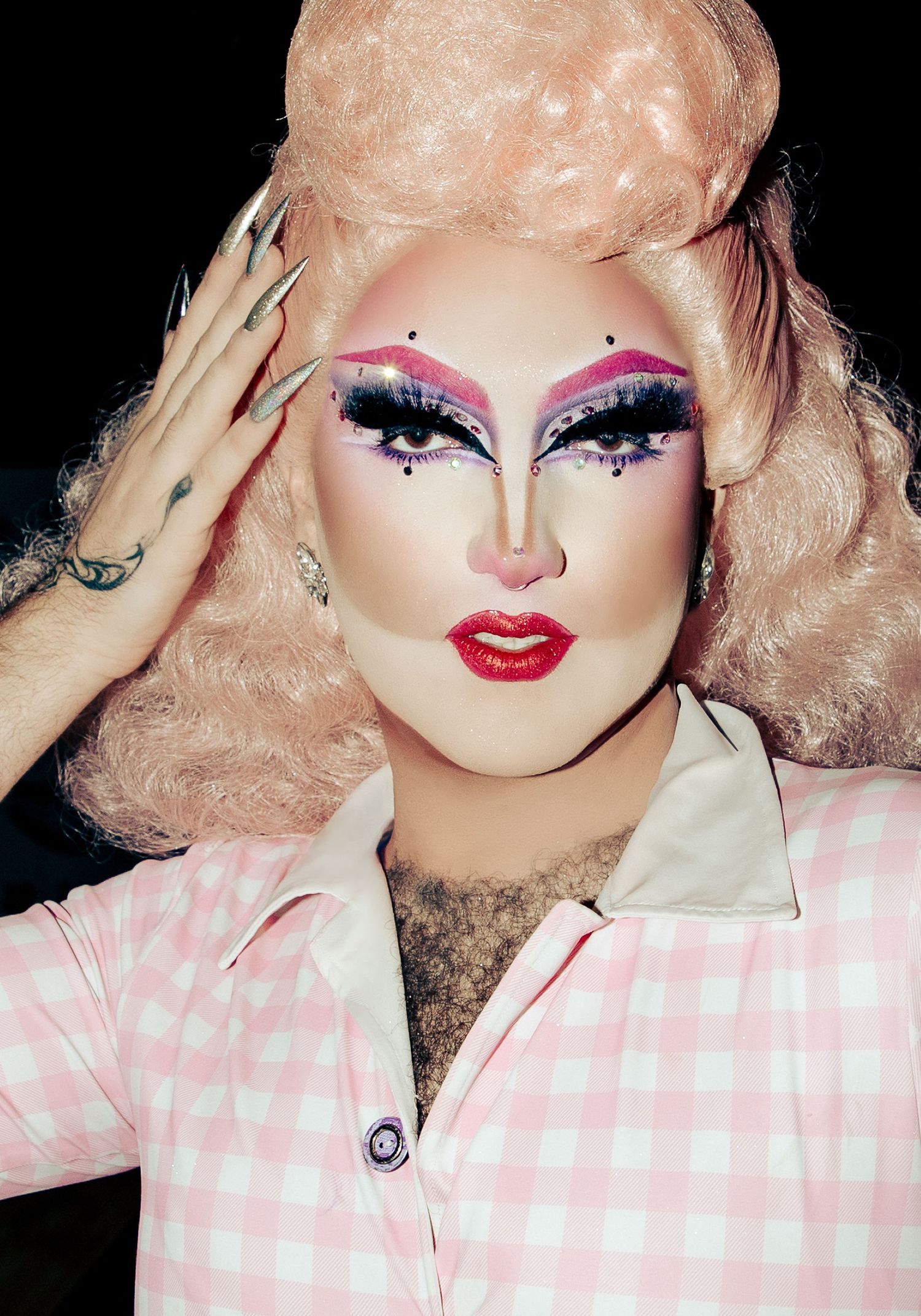 Drag performer, Abork in pink gingham shirt with dramatic eye makeup, jewels, and voluminous blonde updo saluting camera.