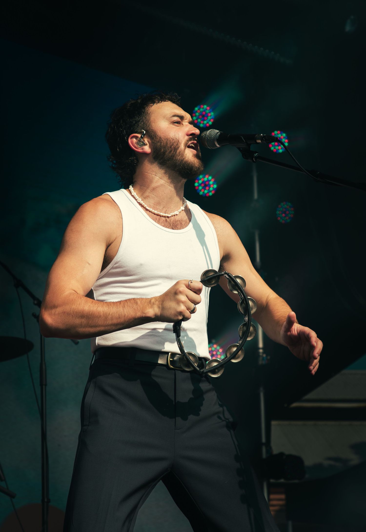 Male lead singer from San Cisco in white tank top performs on stage holding a tambourine with stage lights behind him.